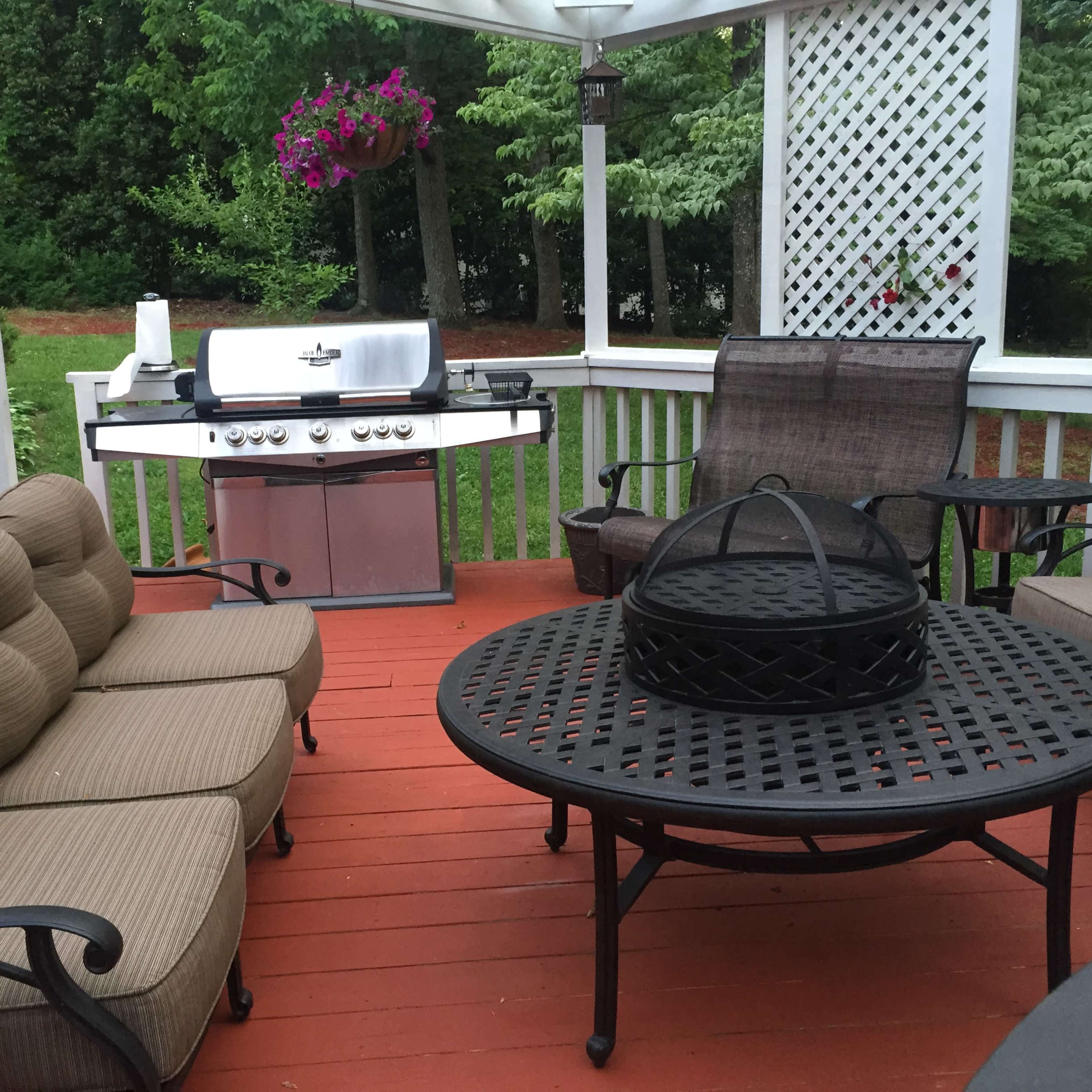 A wooden deck with a grill, a round table, and several cushioned chairs surrounded by green trees.