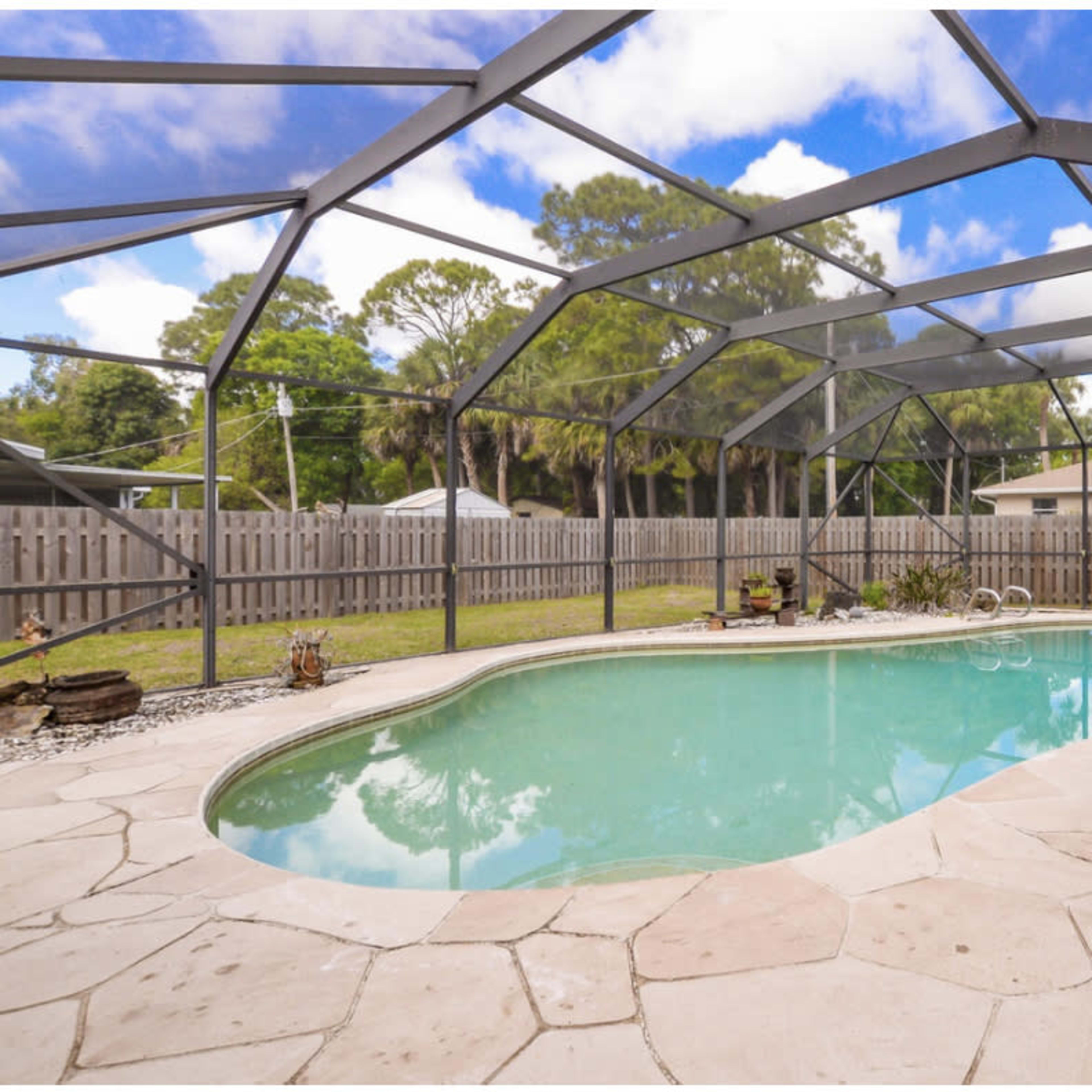 A backyard swimming pool enclosed by a screen enclosure, surrounded by a stone deck and landscaped yard.