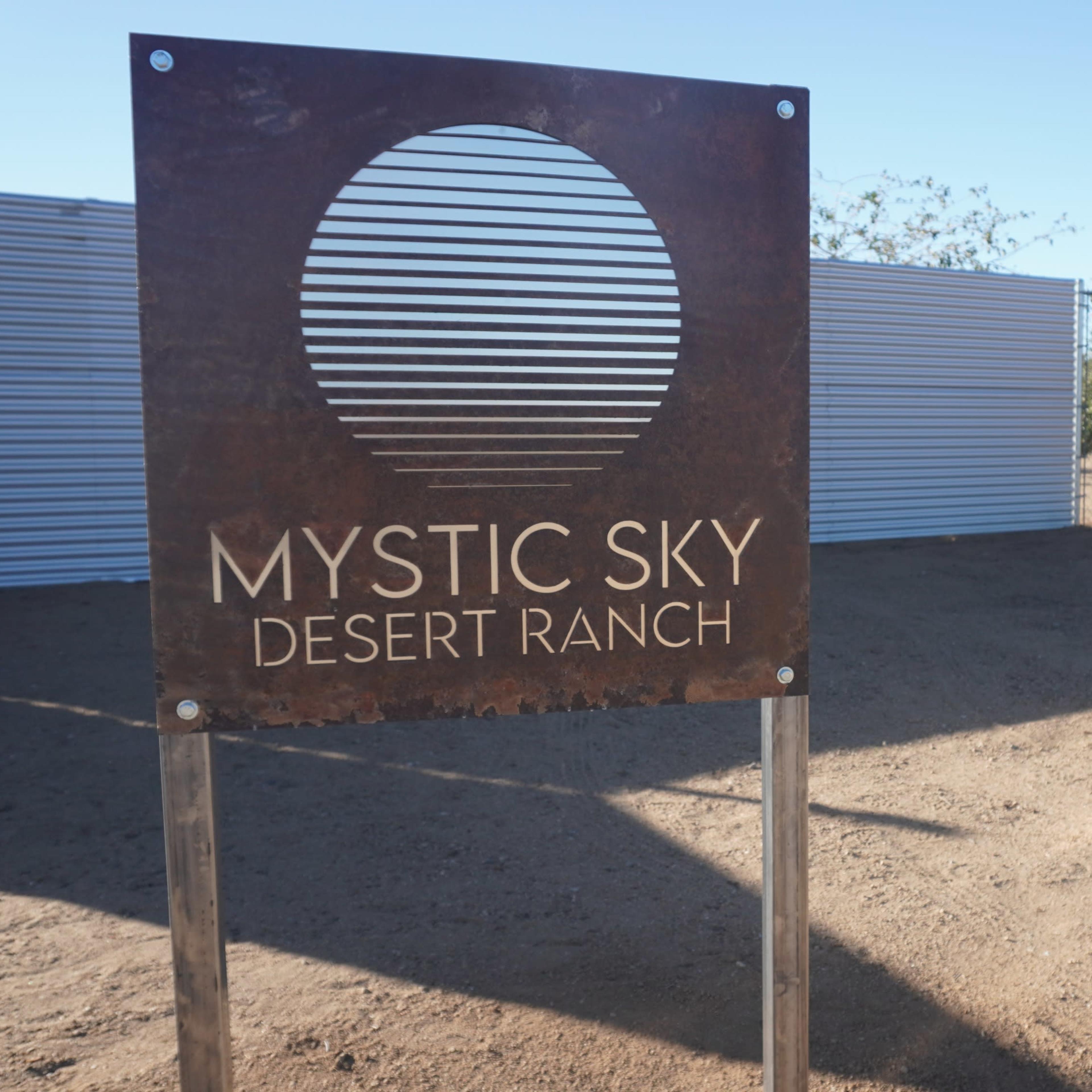 A metal sign for Mystic Sky Desert Ranch stands in a sandy area with sparse vegetation and a clear blue sky in the background.