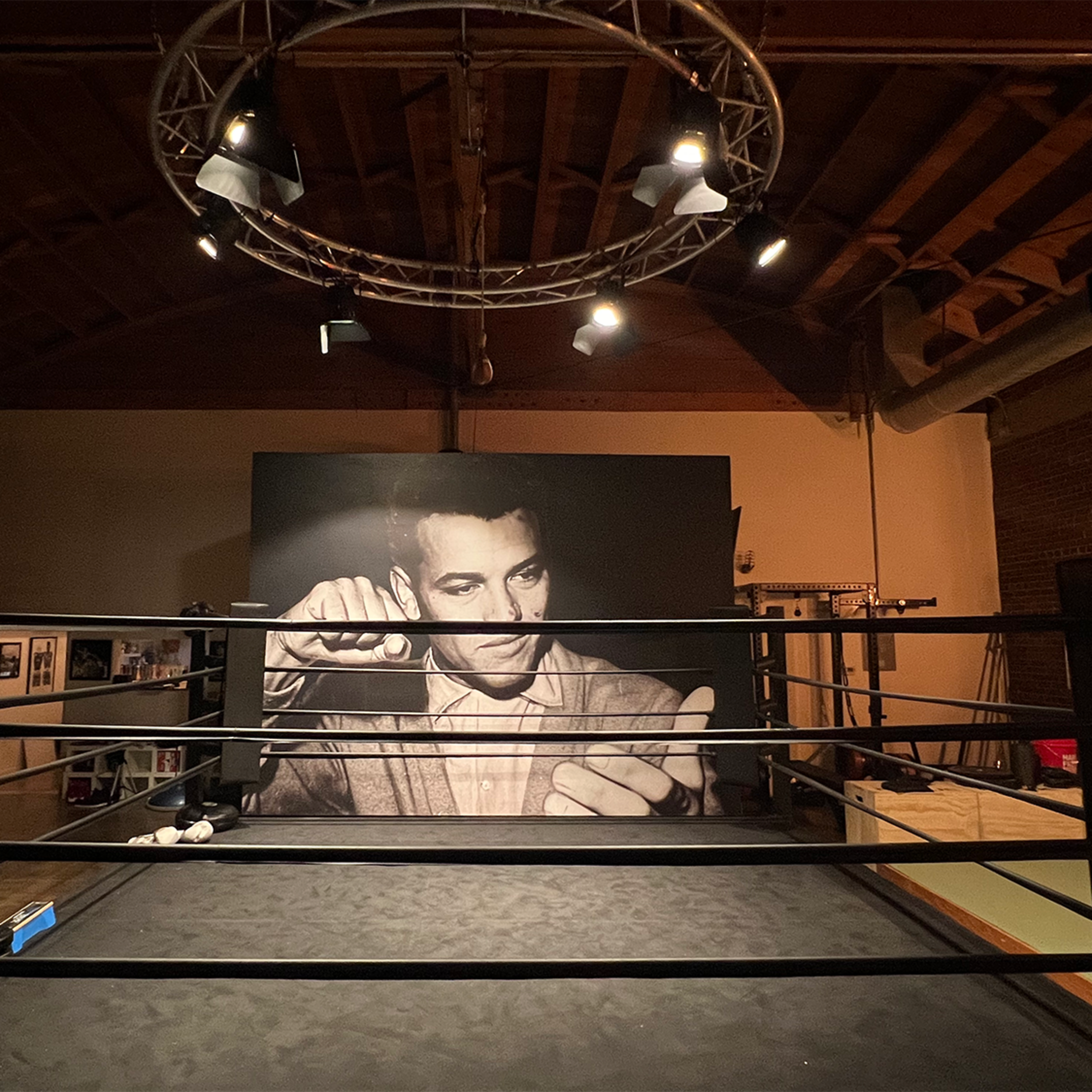 A boxing ring in a dimly lit gym with a large mural of a man in a suit on the wall behind it.