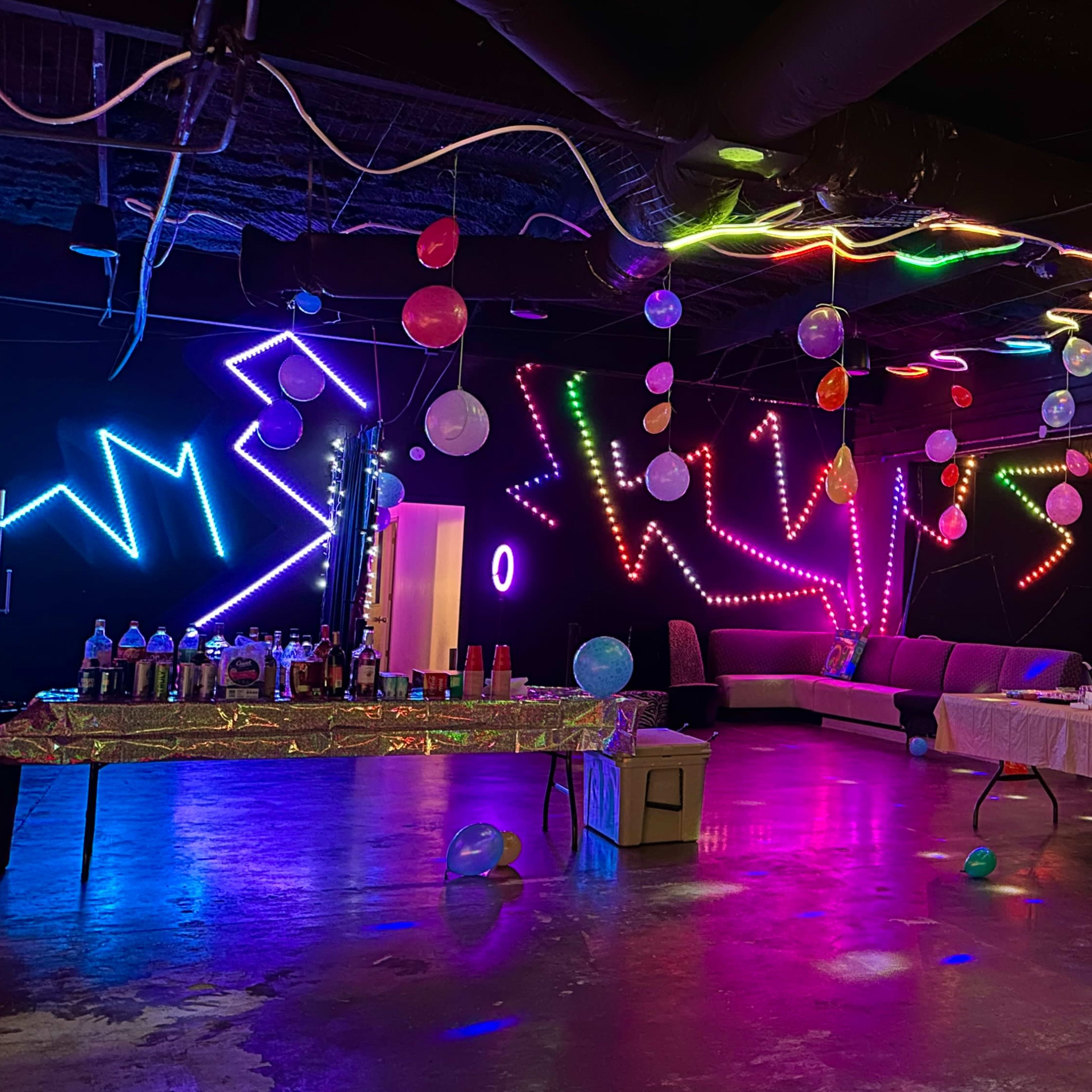 A brightly colored party space features glowing lights, balloons hanging from the ceiling, and tables set up with food and drinks.