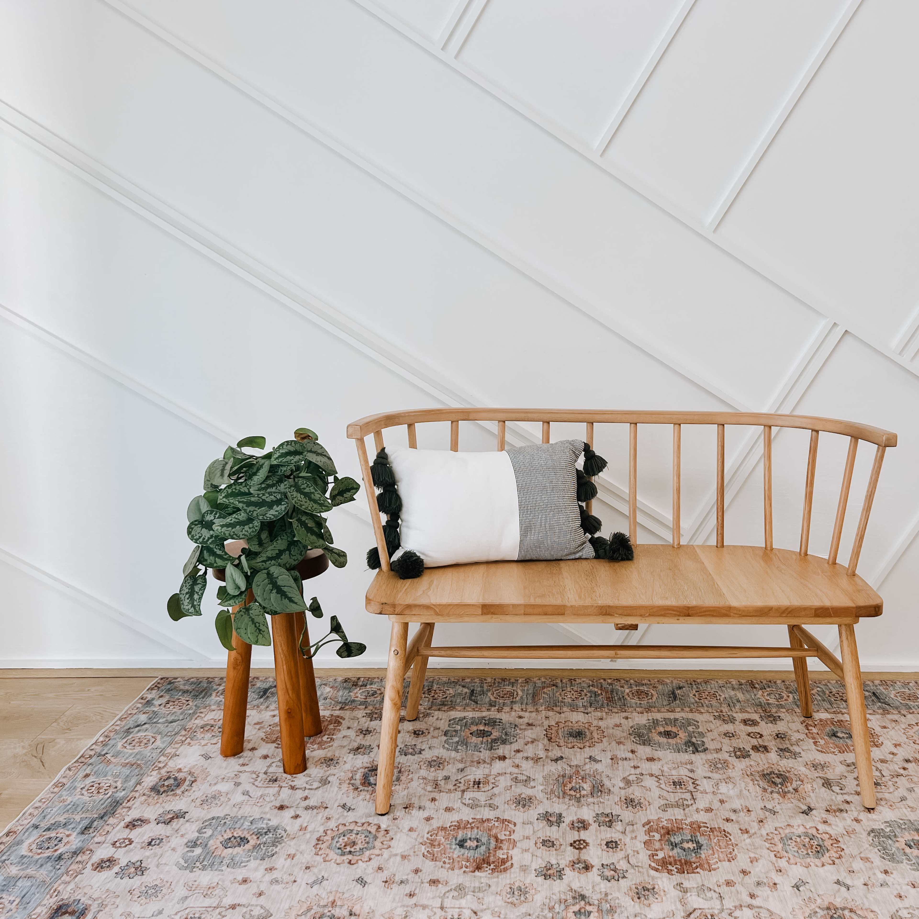 A wooden bench with a cushion sits beside a potted plant on a patterned rug against a diagonal-striped wall.