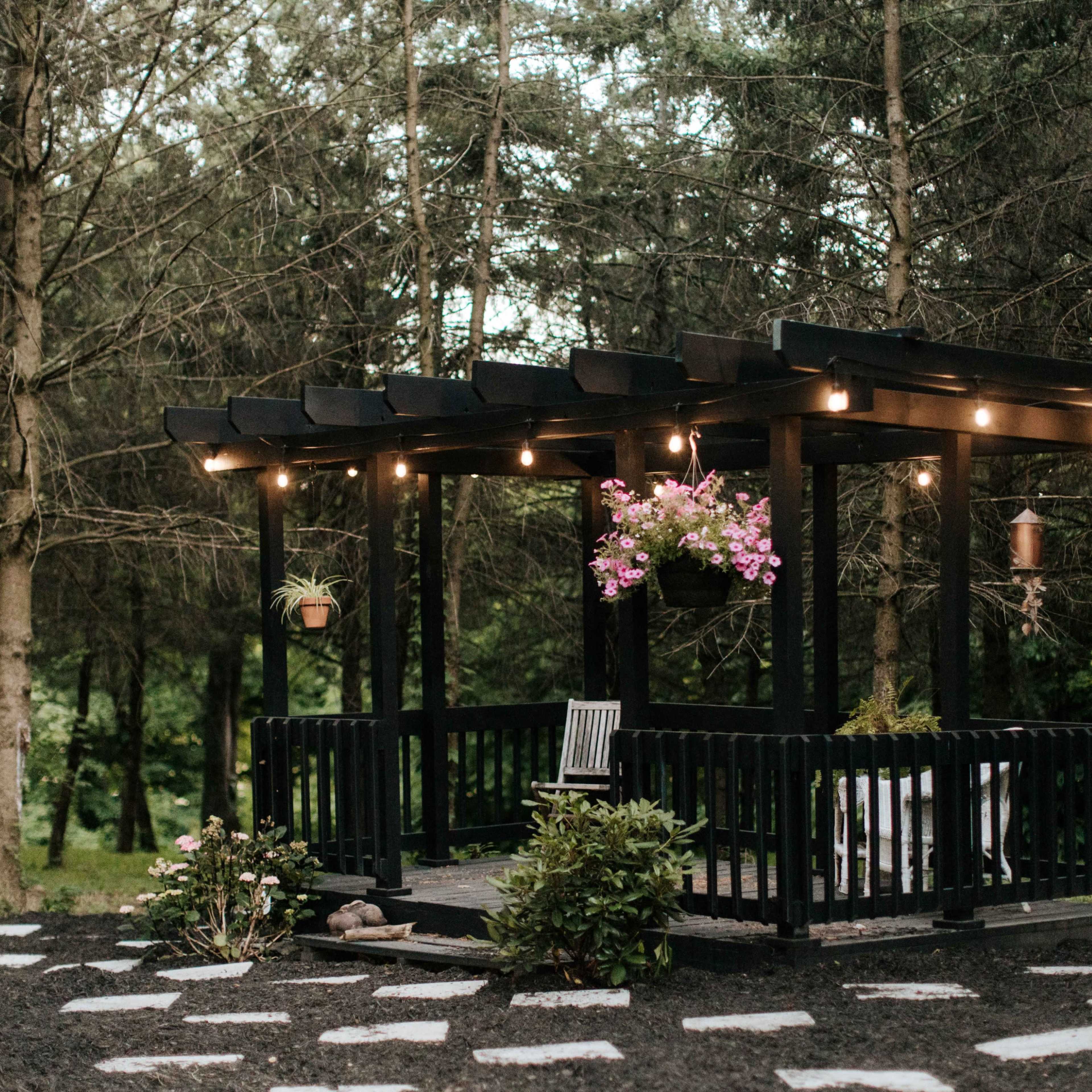 A black wooden gazebo with hanging plants and string lights is situated among trees in a garden area with stone paths.