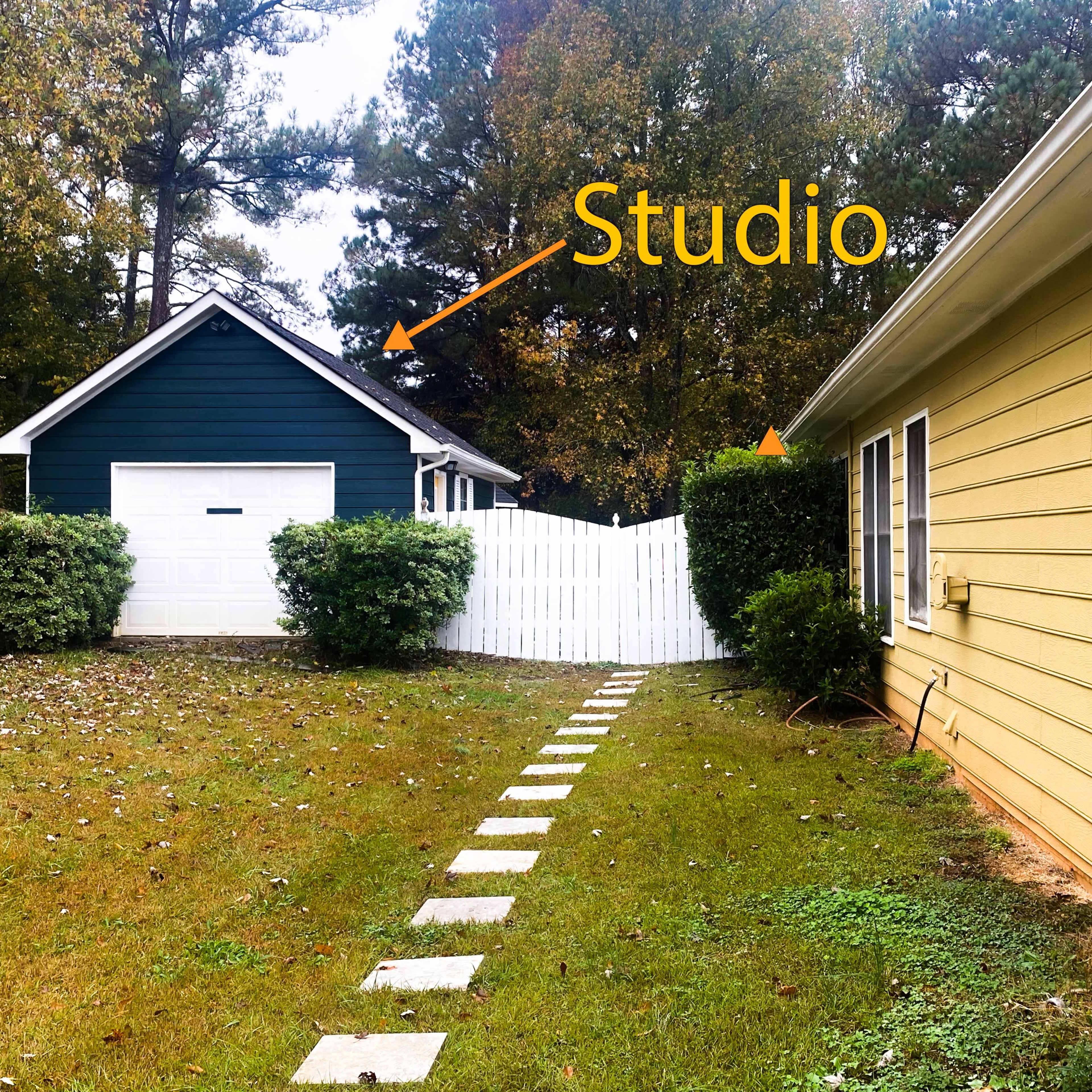 A yellow house adjacent to a green studio building, separated by a white fence and a stone pathway leading through a grassy yard.