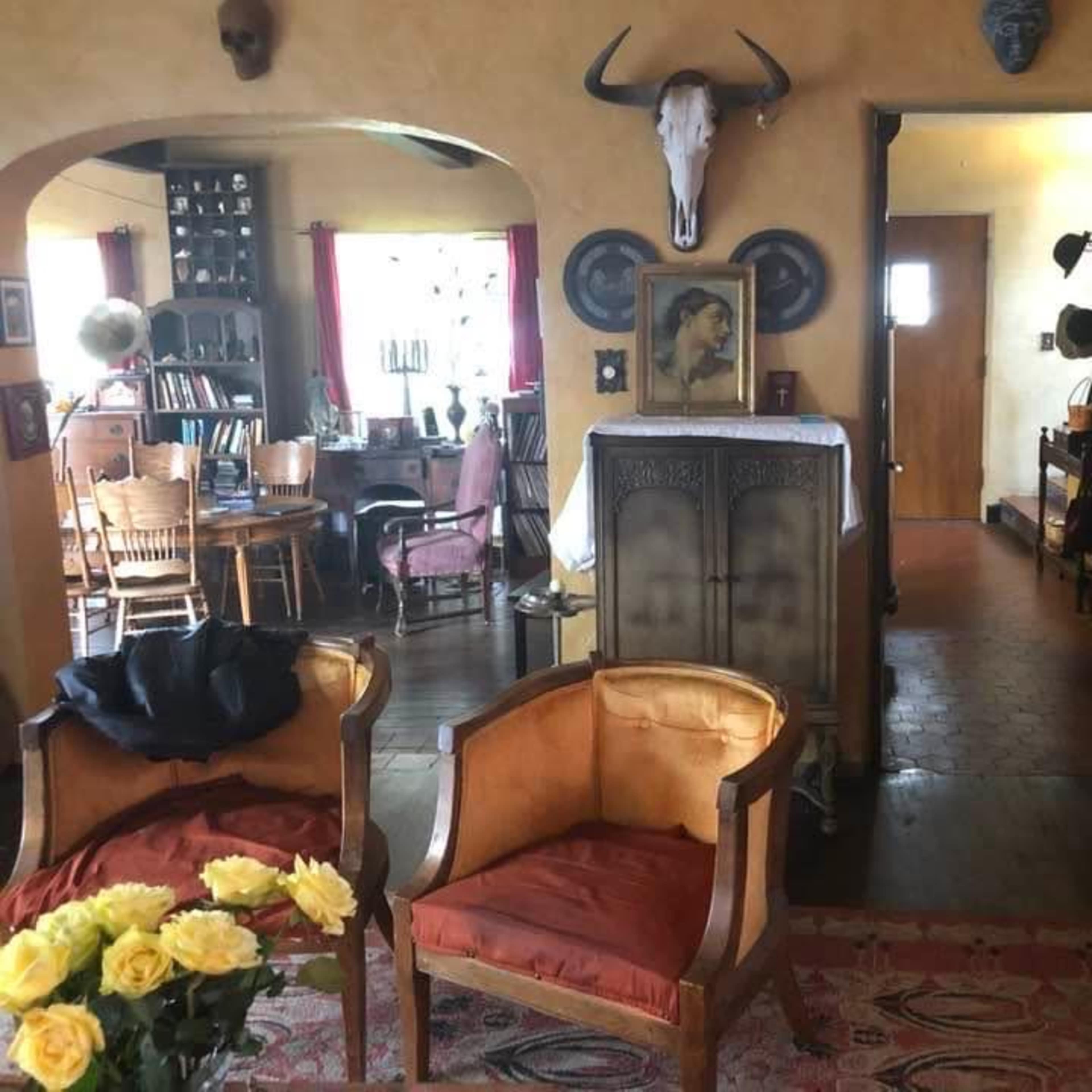 A warmly decorated room featuring two orange armchairs, a wooden dining table with chairs, and a hallway leading to another room, adorned with various wall decorations and a mounted animal skull.