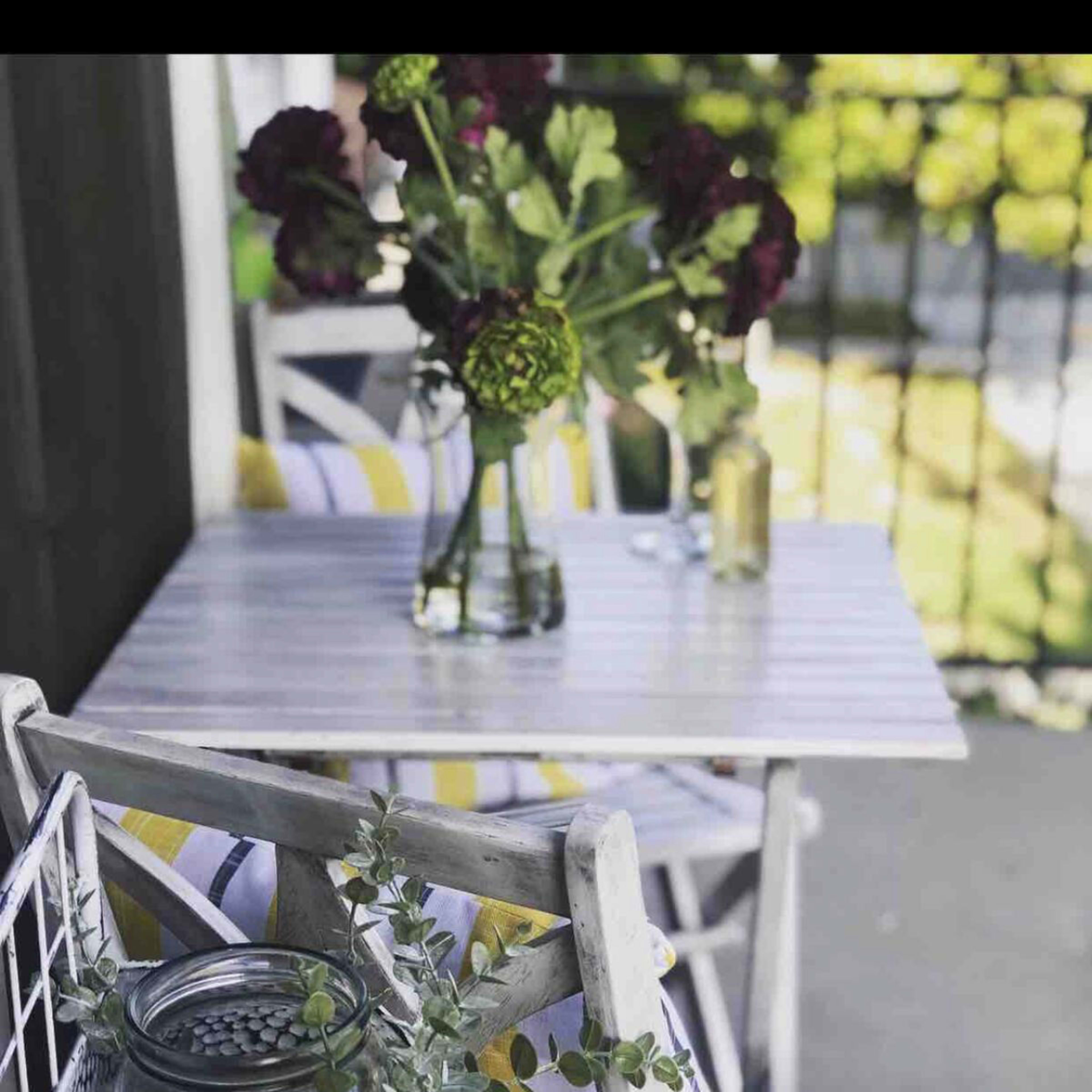 A small, white-striped table is set on a porch, adorned with a vase of flowers and accompanied by colorful chairs and a glass jar.