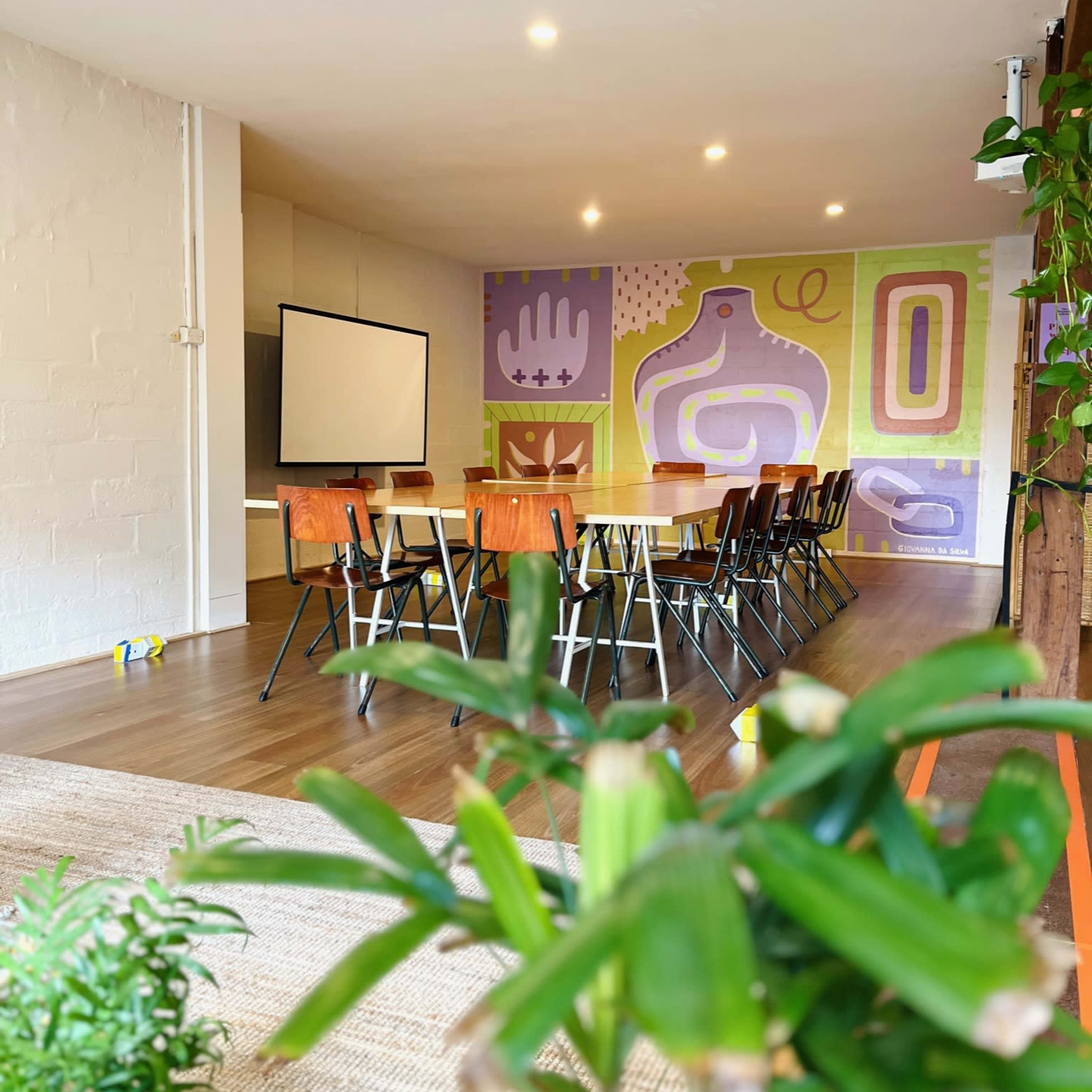 A spacious meeting room features a long table surrounded by chairs, with a colorful mural on the wall and a projector screen at the front.