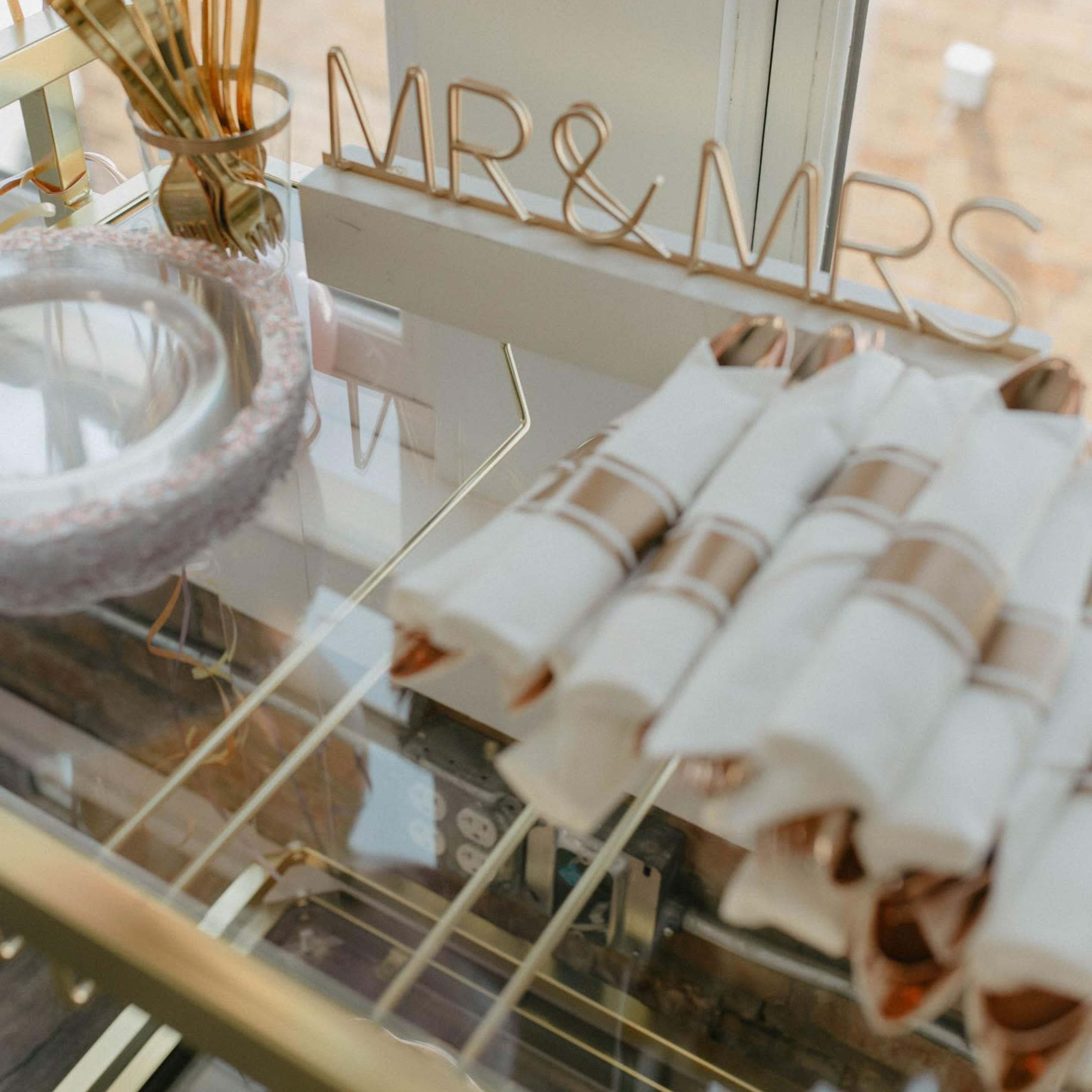 A glass display case features a "MR & MRS" sign alongside neatly rolled white napkins and gold utensils.