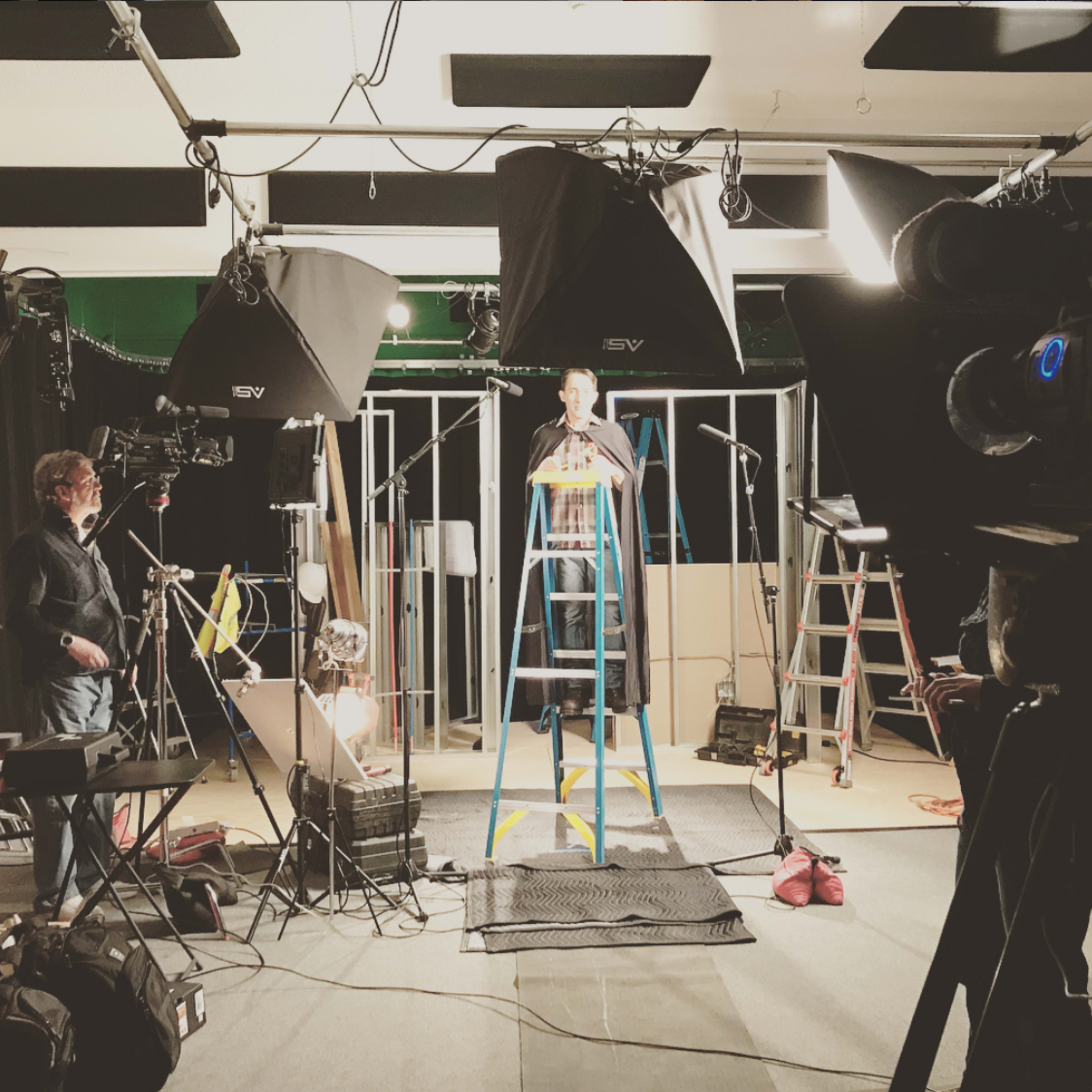 A man stands on a ladder in a studio surrounded by cameras, lights, and various equipment.