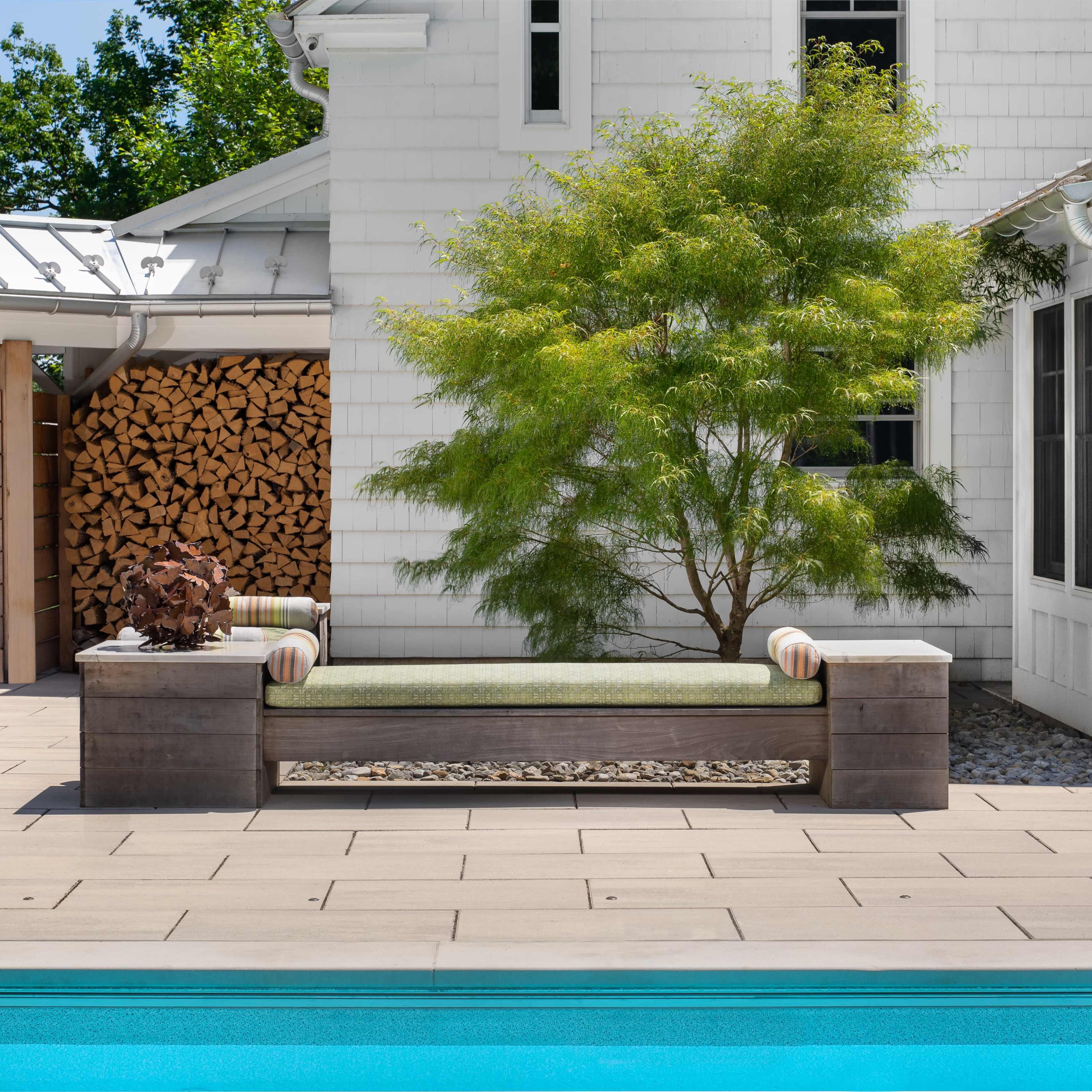 A wooden bench with green cushions is positioned beside a pool, framed by a small tree and a woodstack in the background.