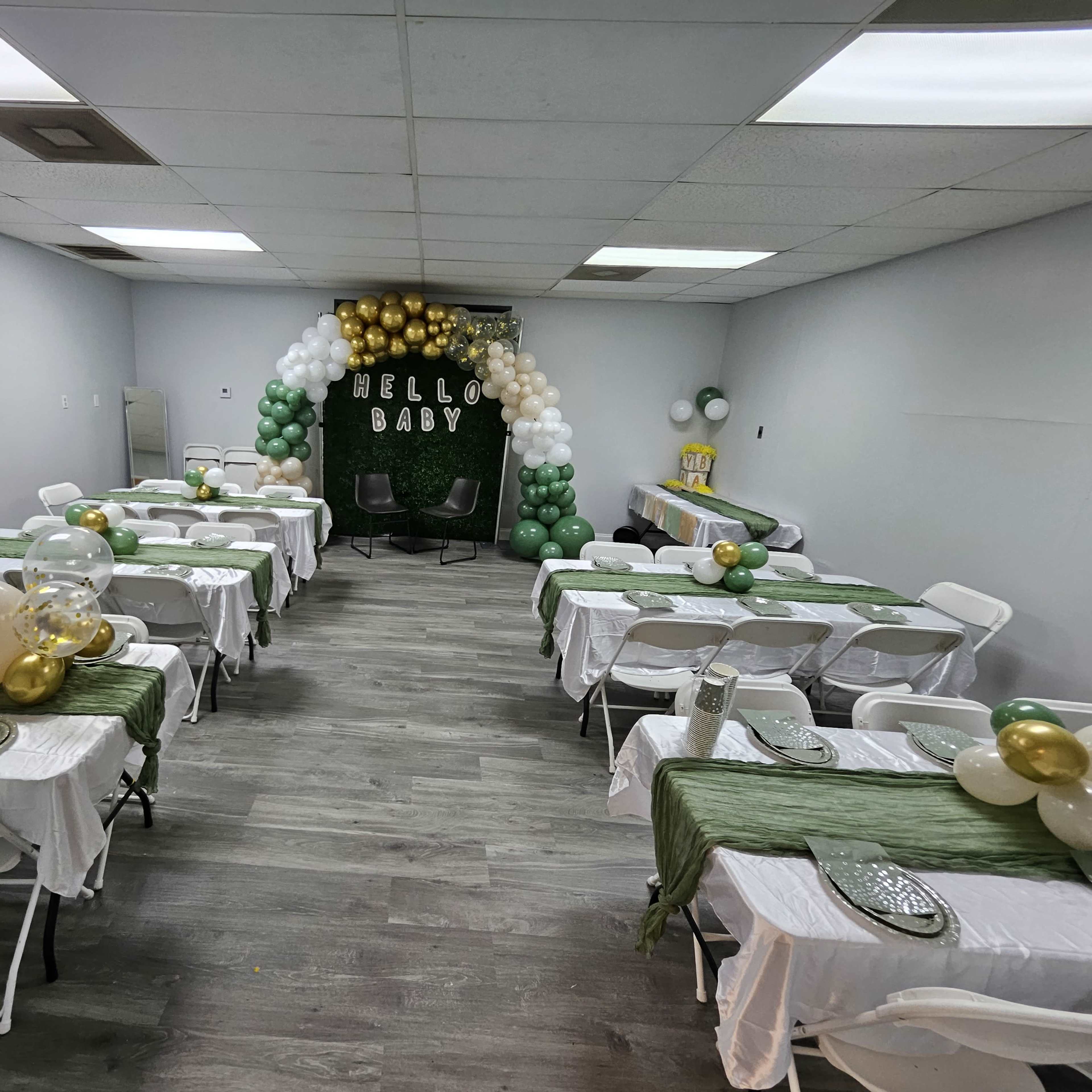 A decorated room for a baby shower, featuring several tables with white tablecloths, green and gold balloons, and a backdrop that reads "HELLO BABY."