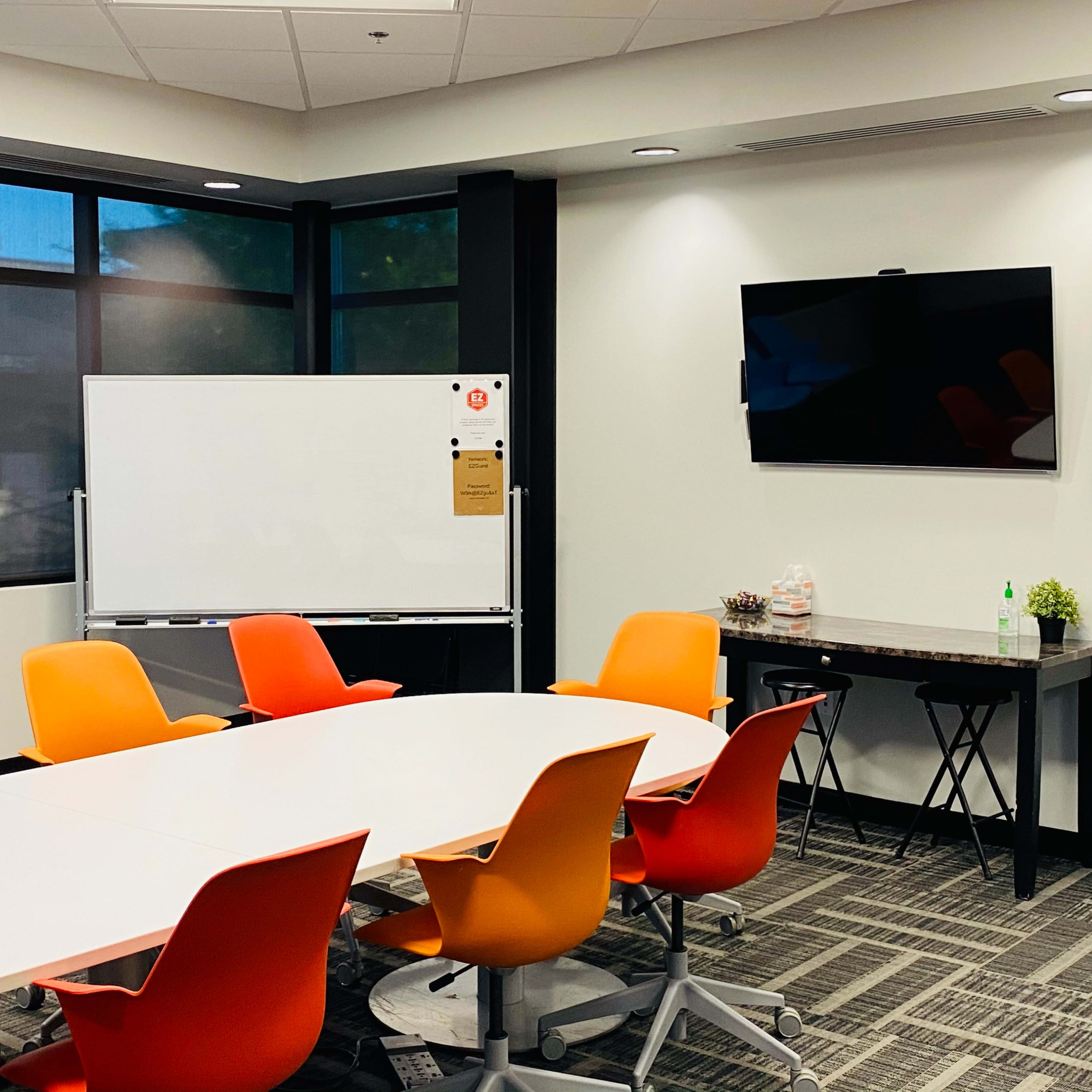 A brightly lit conference room features a large whiteboard, a television mounted on the wall, and a table surrounded by colorful chairs.