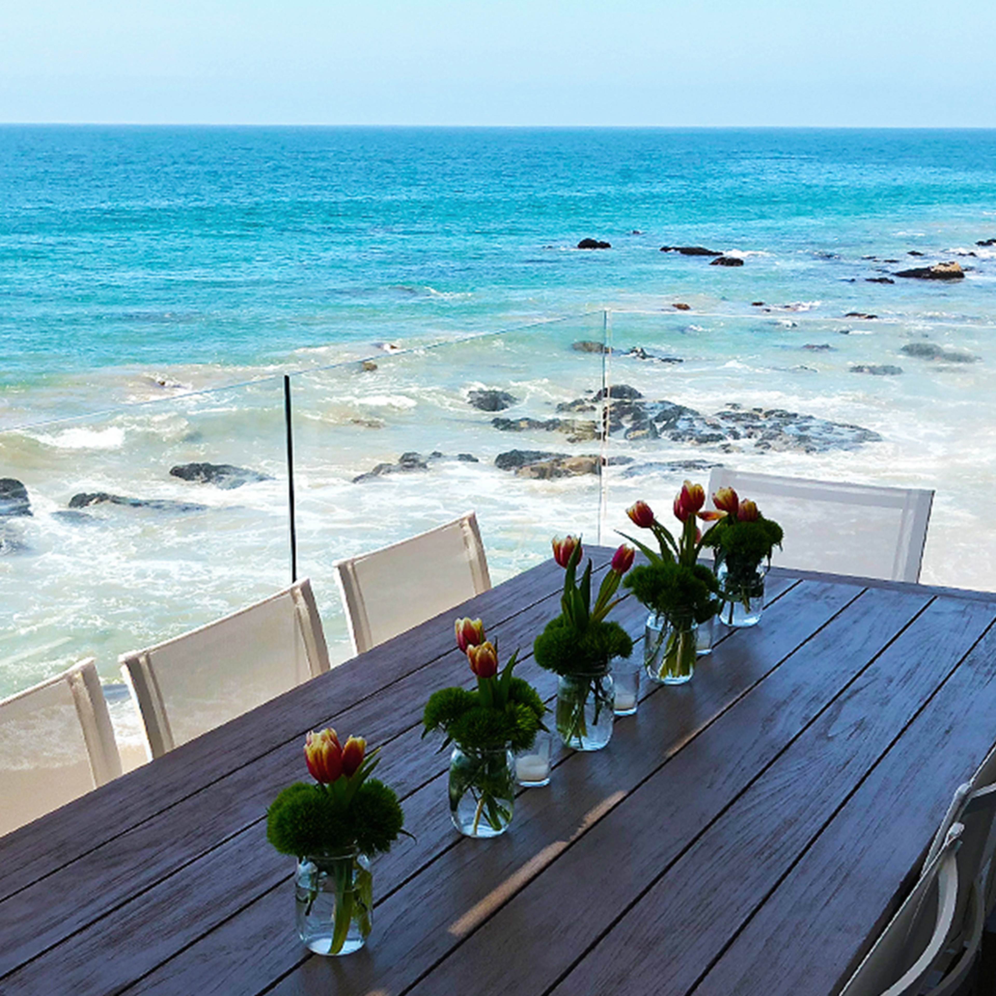 A seaside table is set with vases of flowers, overlooking the ocean and rocky shore.