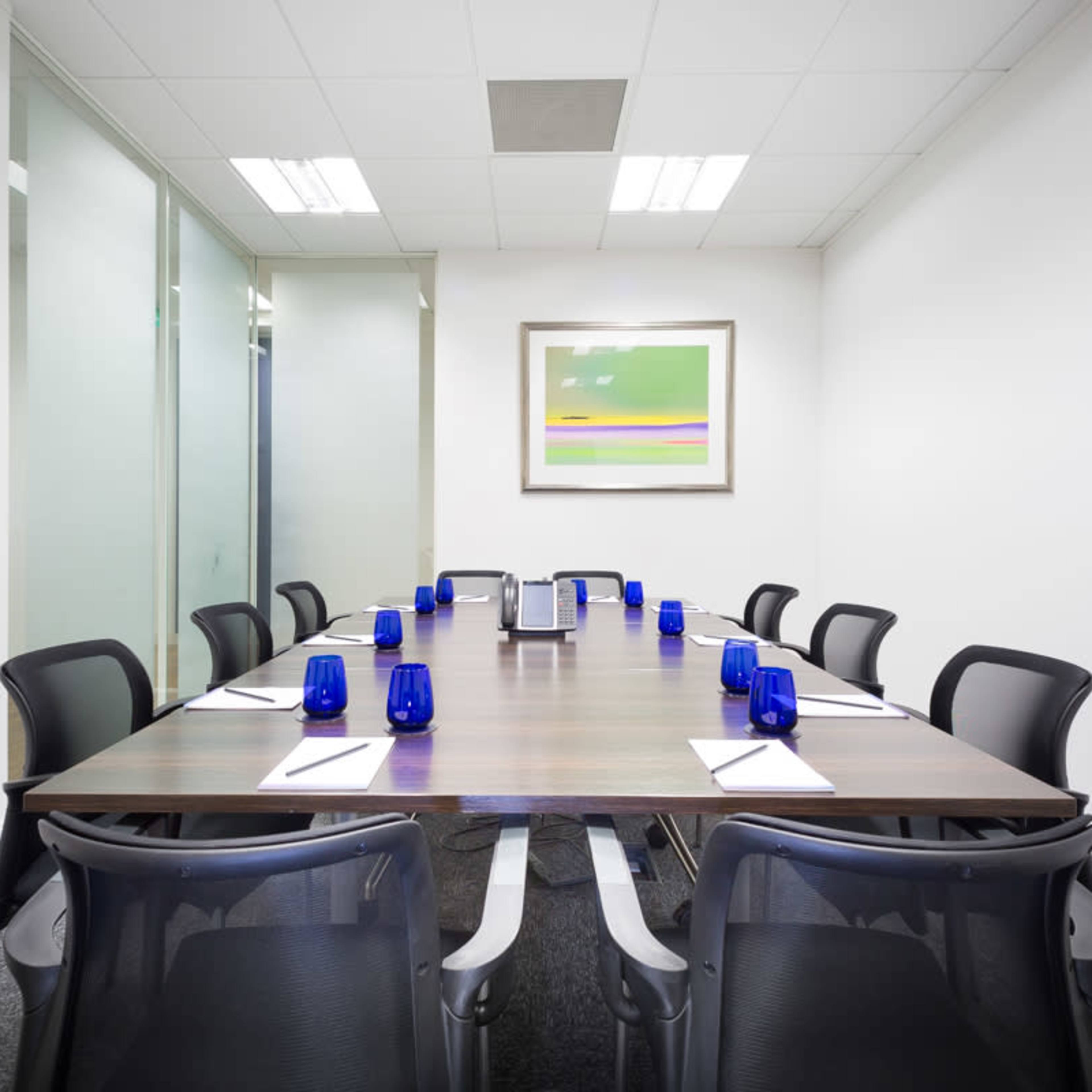 A long wooden conference table is set with blue glasses and notepads, surrounded by black chairs in a brightly lit meeting room.
