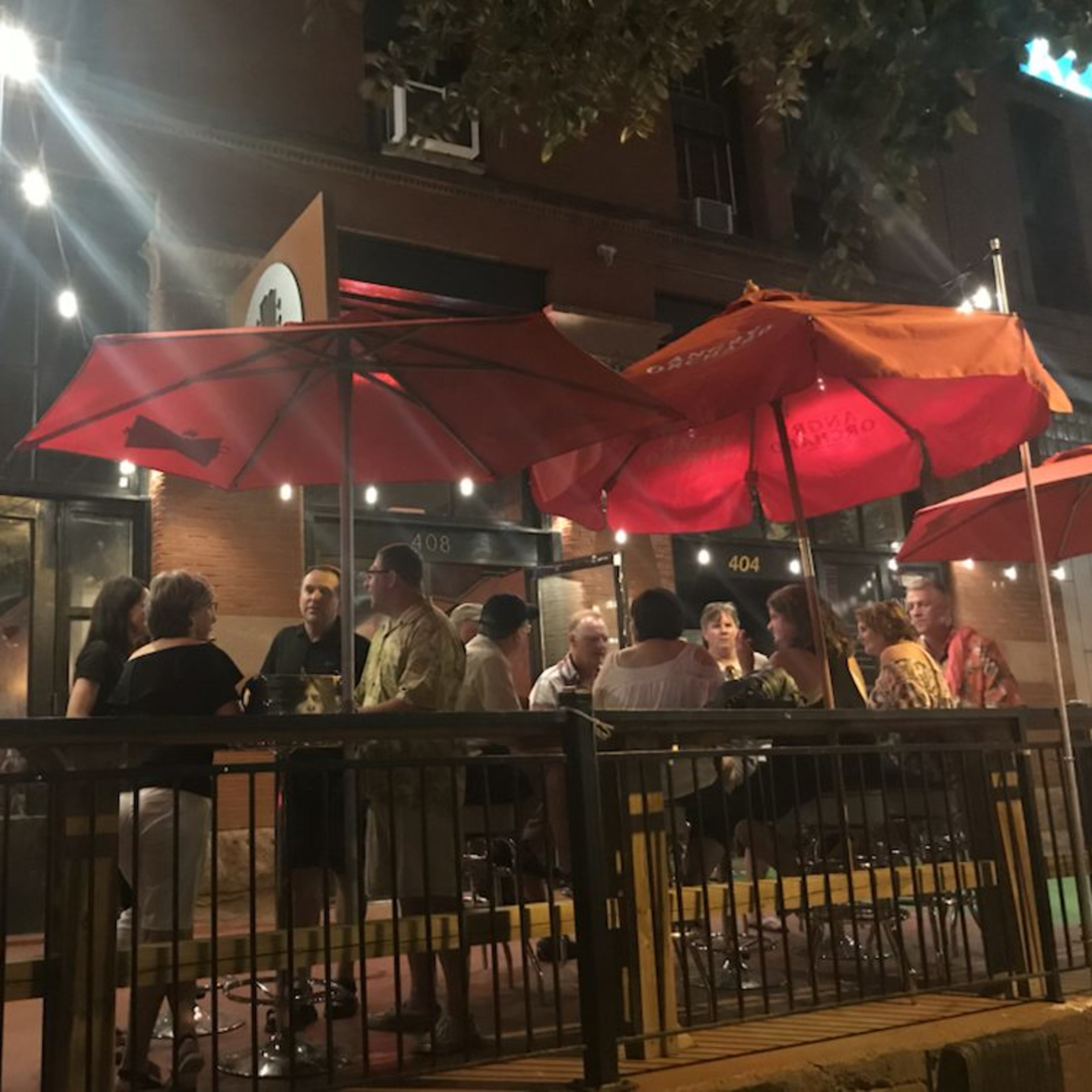 A group of people gathers around tables with red umbrellas outside a restaurant at night.
