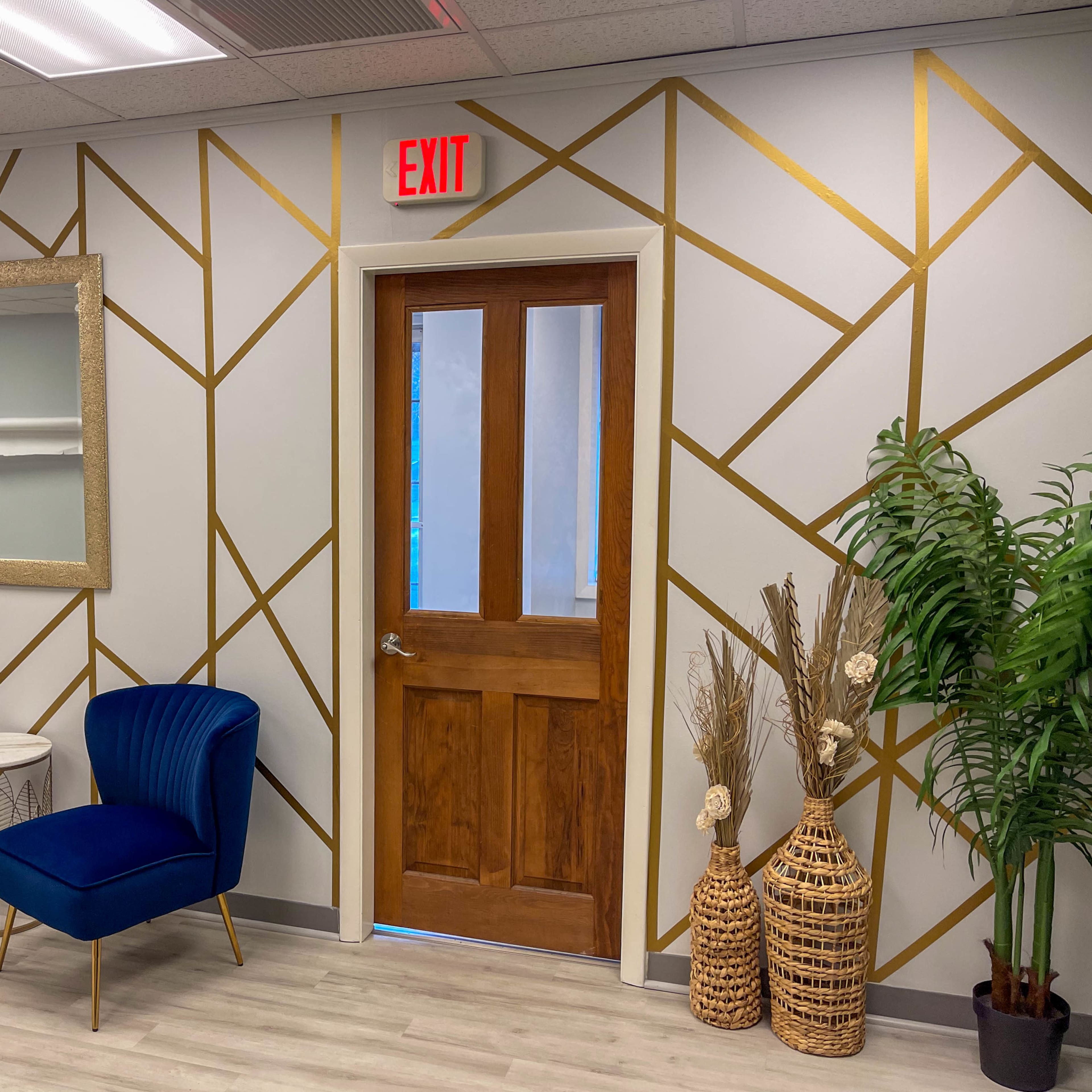 A modern waiting area with blue upholstered chairs, a decorative mirror, and a door framed by geometric gold lines on the wall.