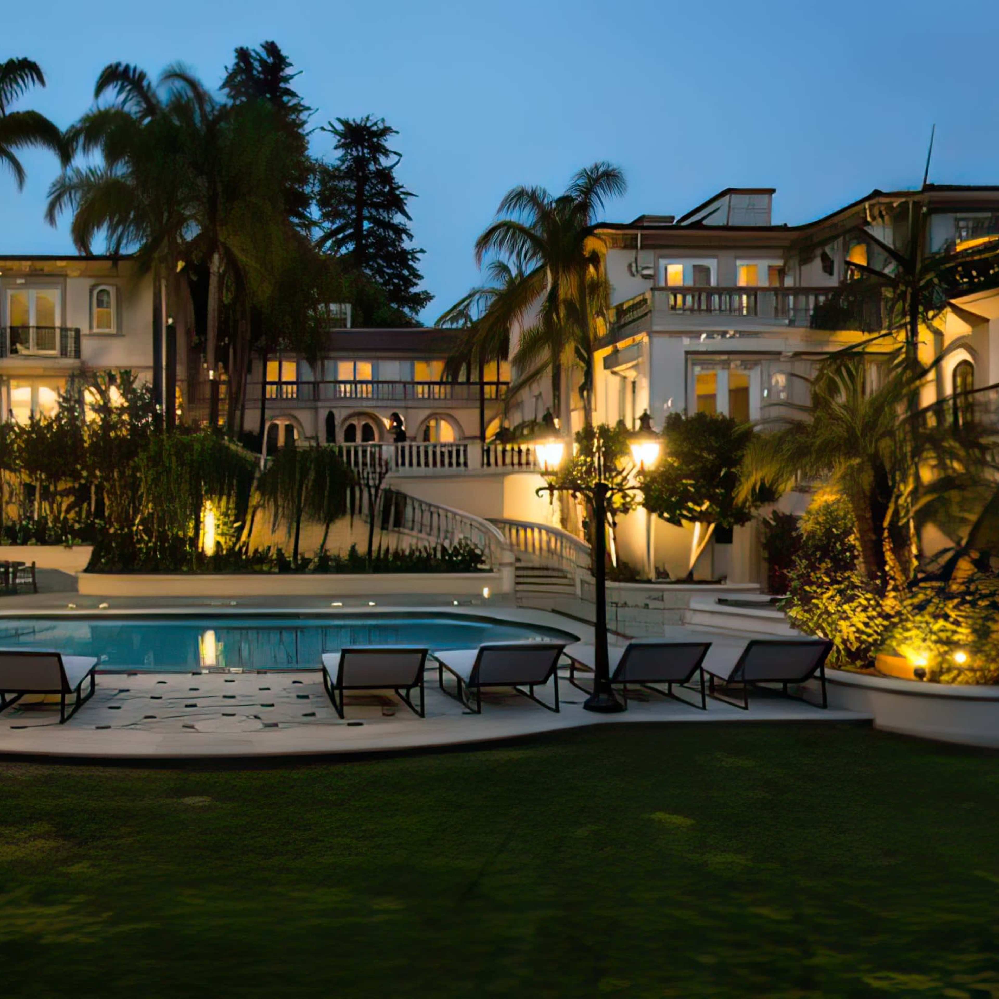 A spacious mansion with a swimming pool is surrounded by palm trees and illuminated by outdoor lighting at dusk.