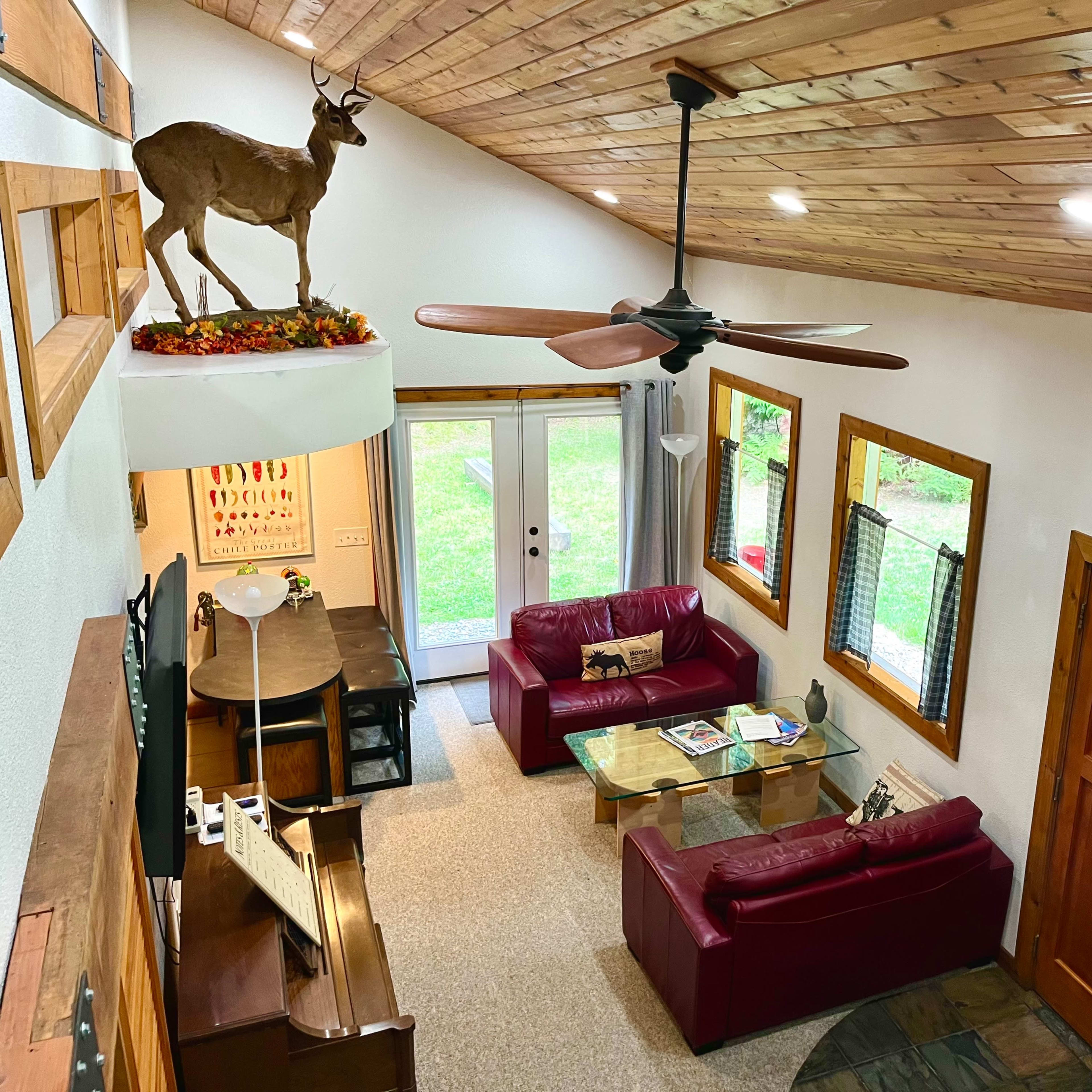 A cozy living room with red couches, a wooden dining table, and a mounted deer head decoratively placed above a door, all situated beneath a vaulted wooden ceiling.