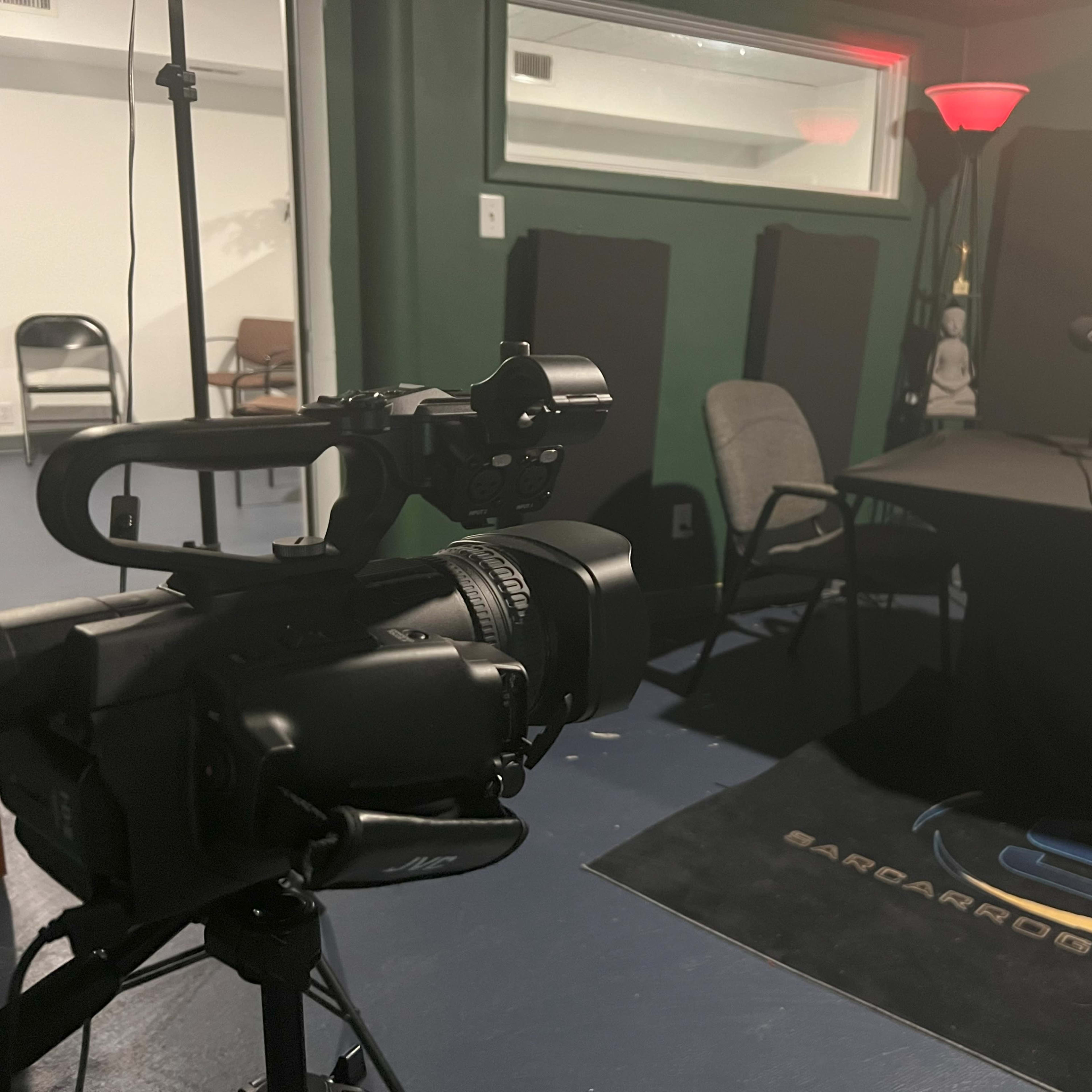 A video camera focused on a table set up with microphones and chairs in a recording studio.