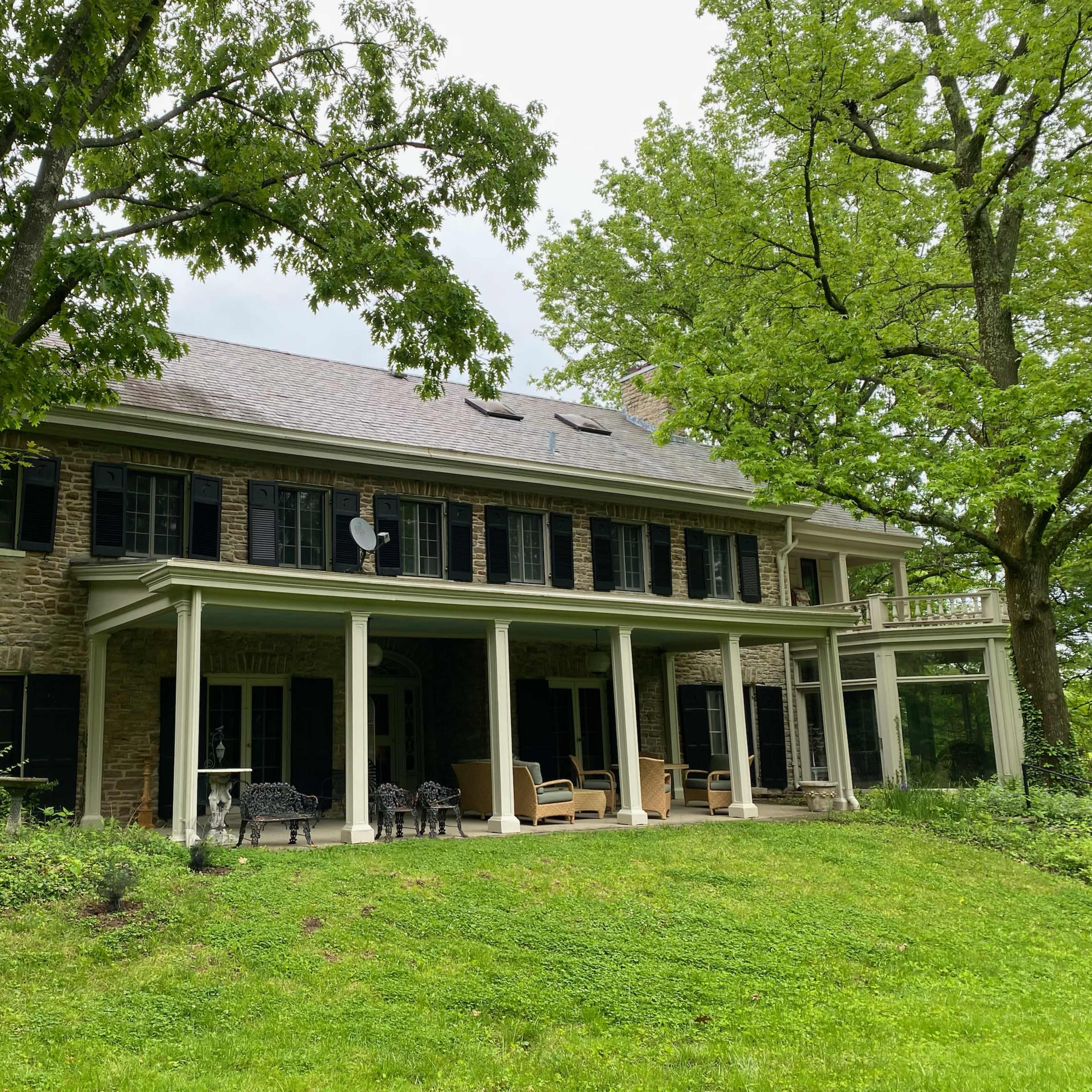 A large two-story stone house with dark shutters and a covered porch, set among green trees and grass.