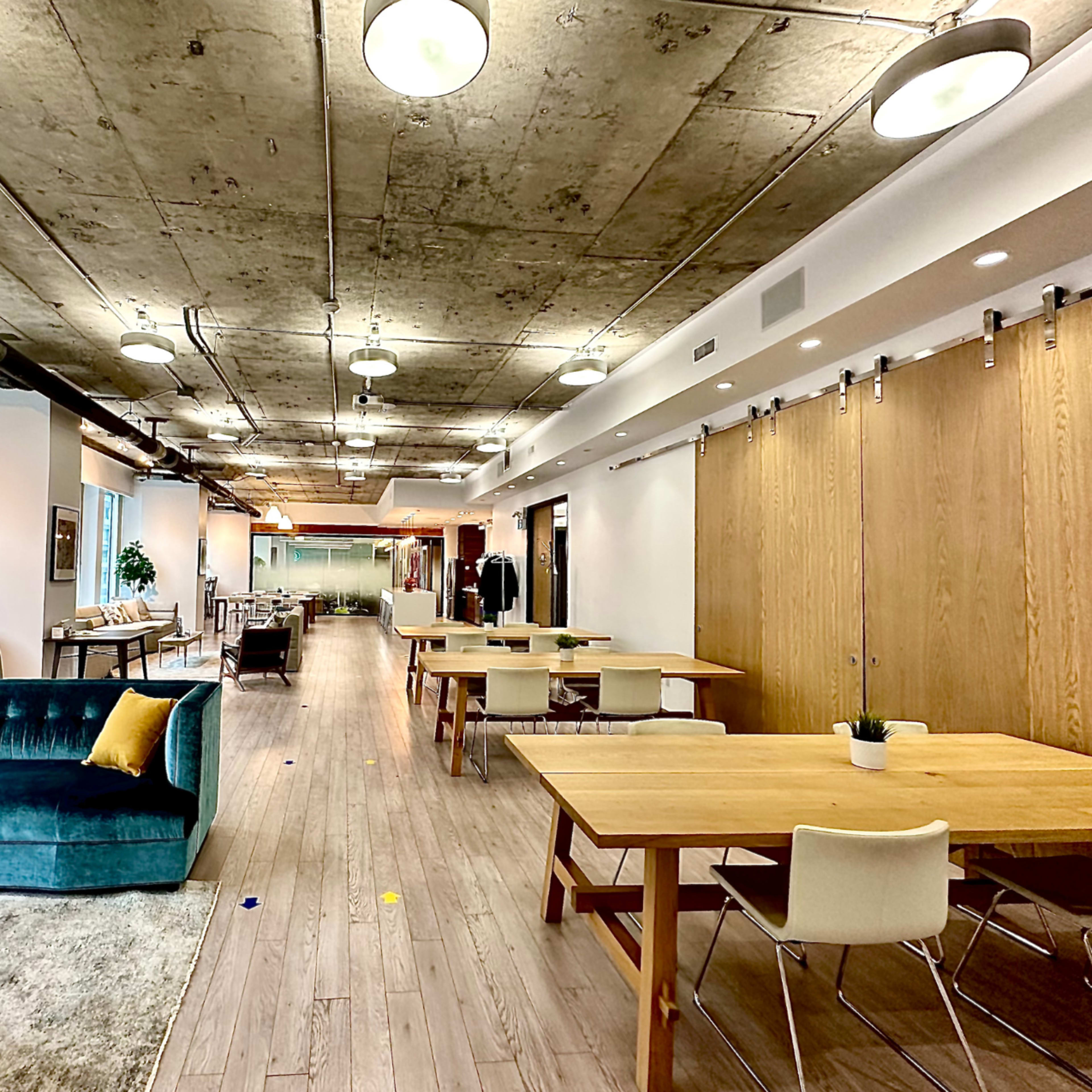 A modern office space with a long hallway featuring wooden tables, a blue couch, and large windows.