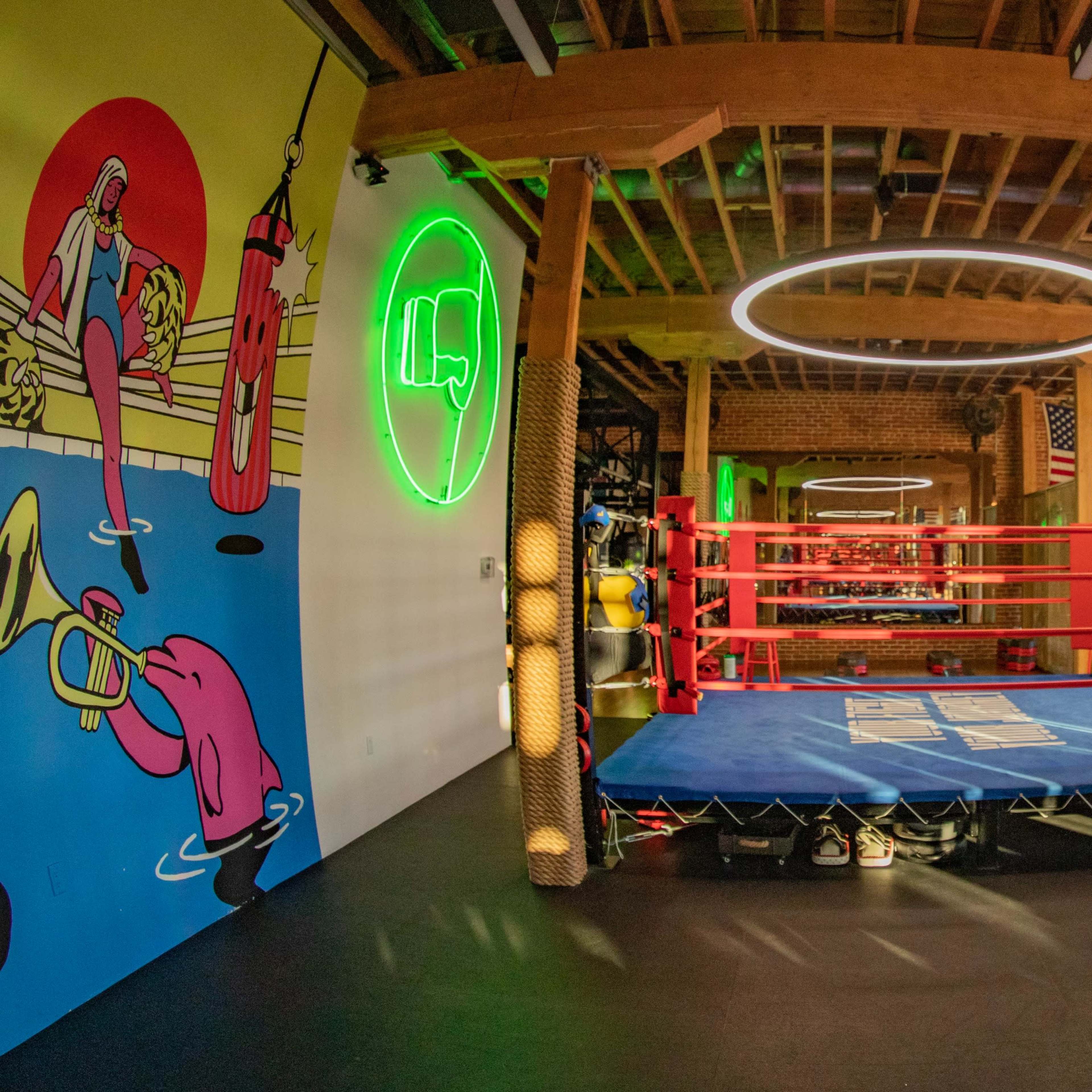 A boxing gym features a central ring surrounded by vibrant wall art, seating, and fitness equipment.