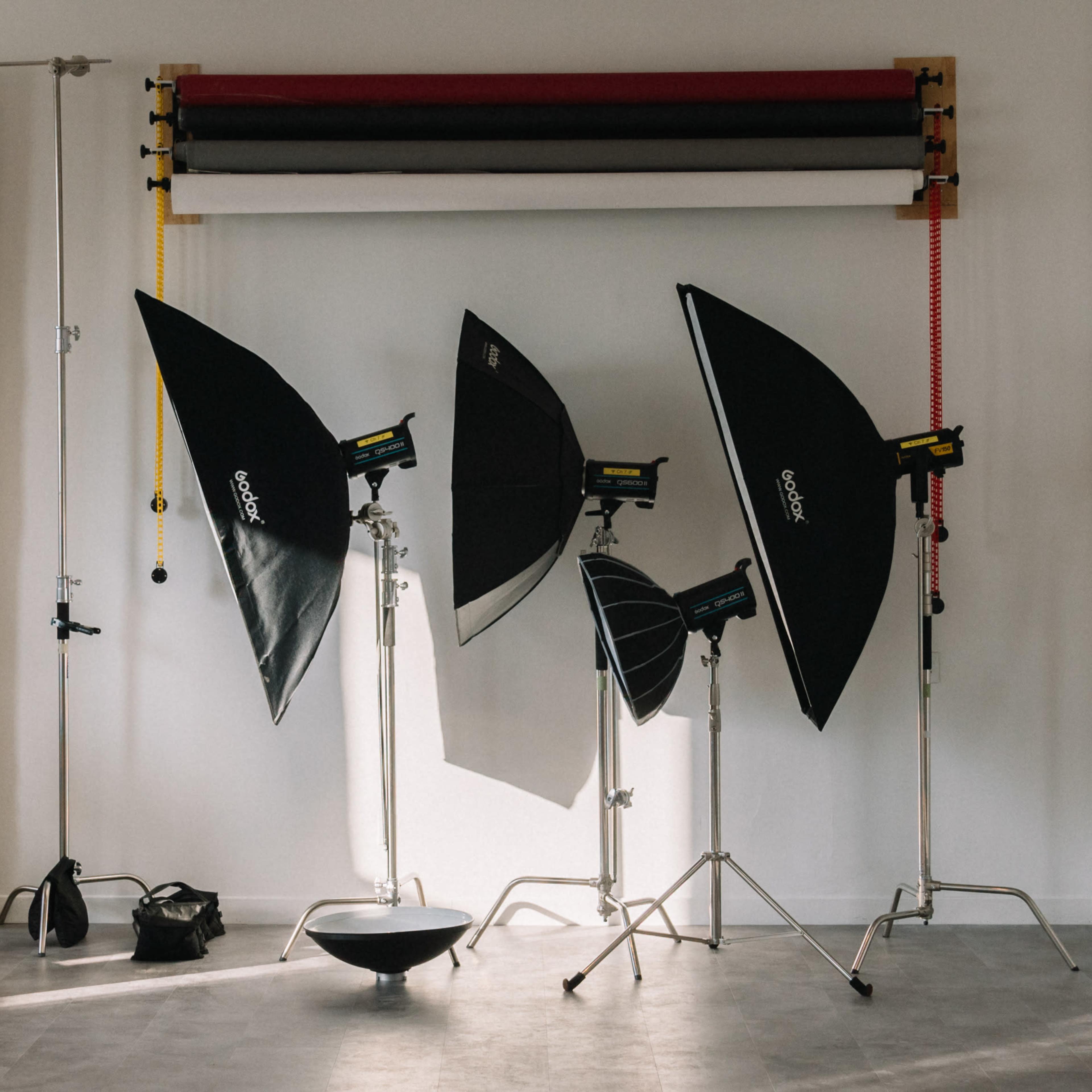 A photography studio setup with several softbox lights, a backdrop roller, and various equipment arranged against a wall.