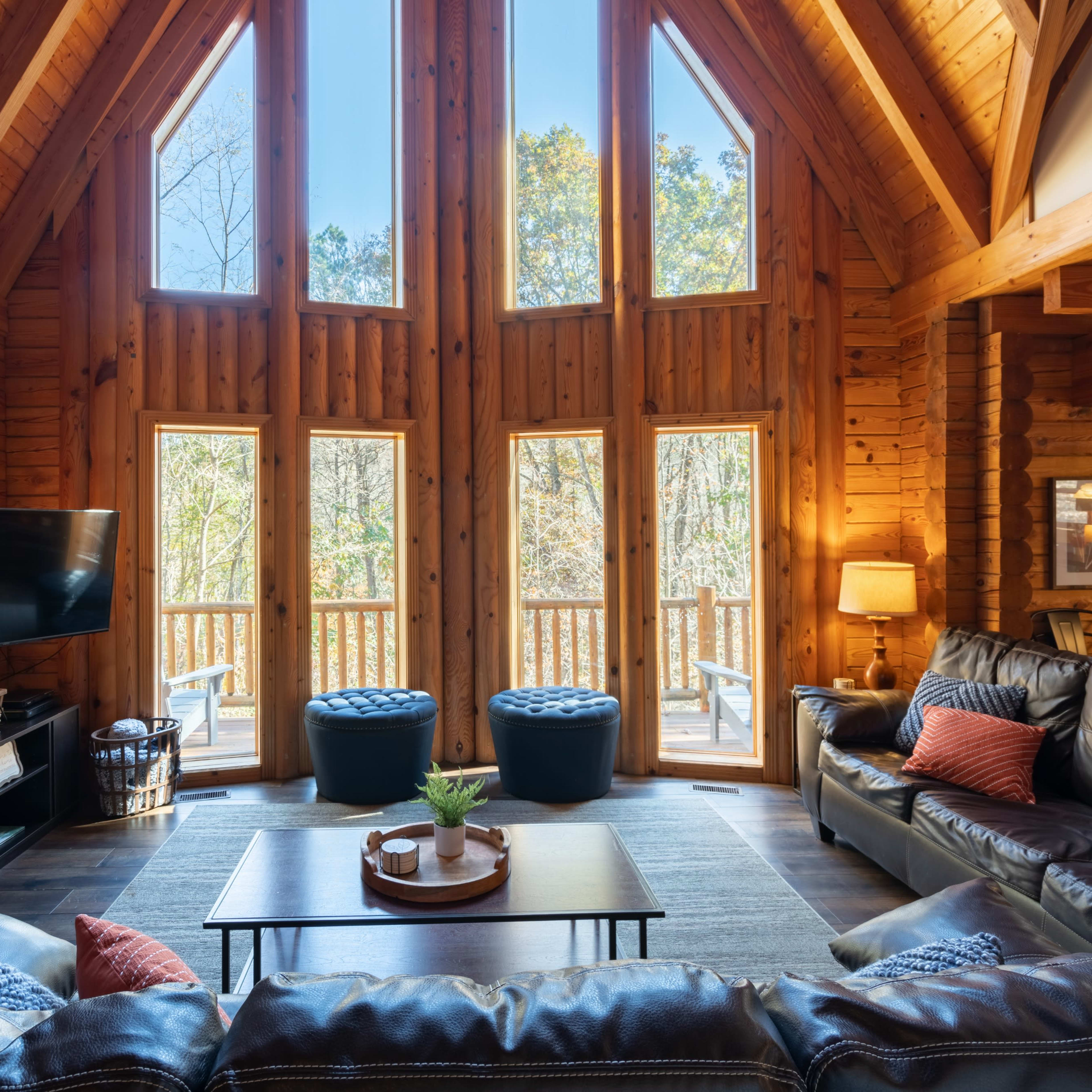 The living room features large windows that frame a view of trees, wooden walls, a stone fireplace, and leather seating arrangements.