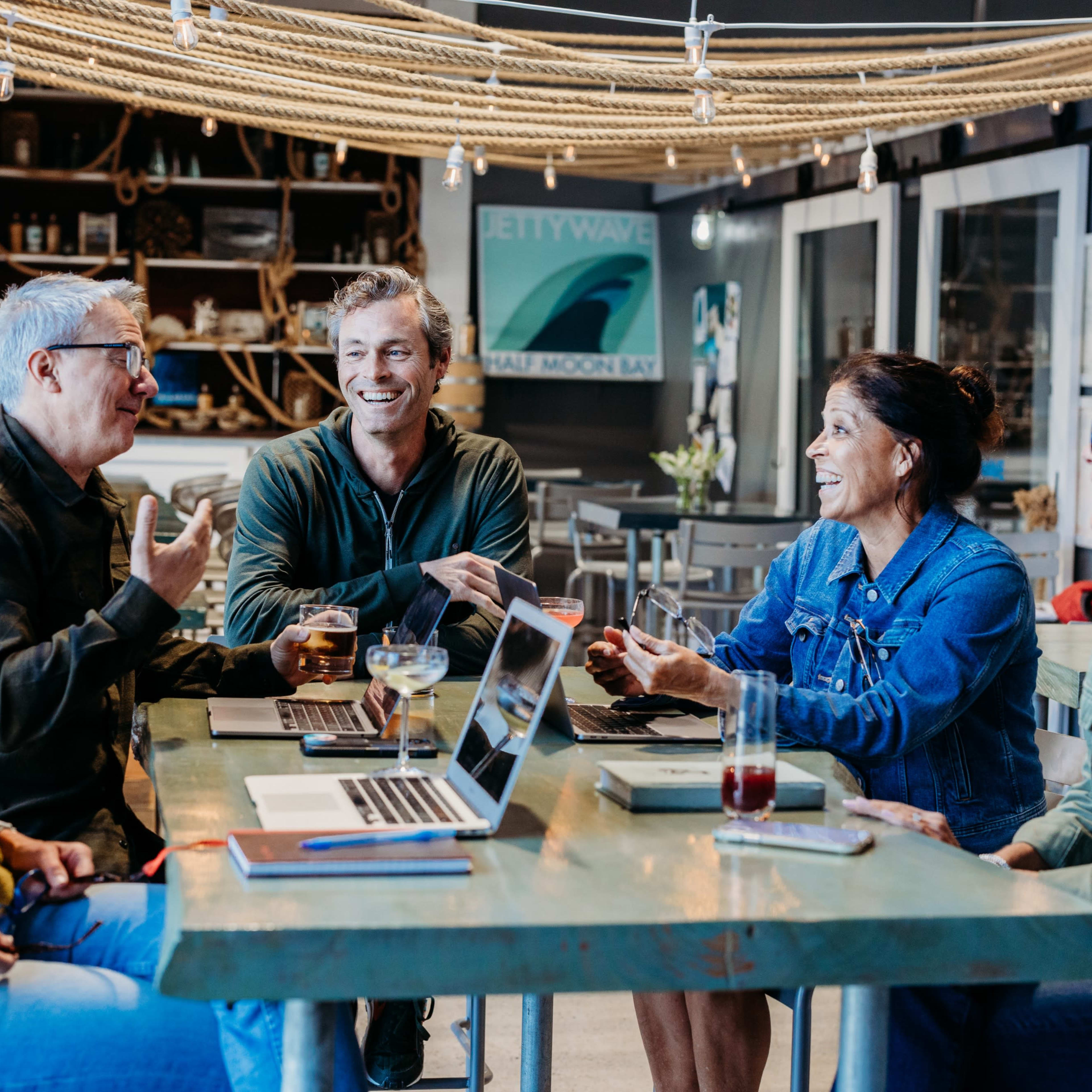 A group of six people engages in a discussion around a table with laptops and drinks in a cozy, well-lit café.