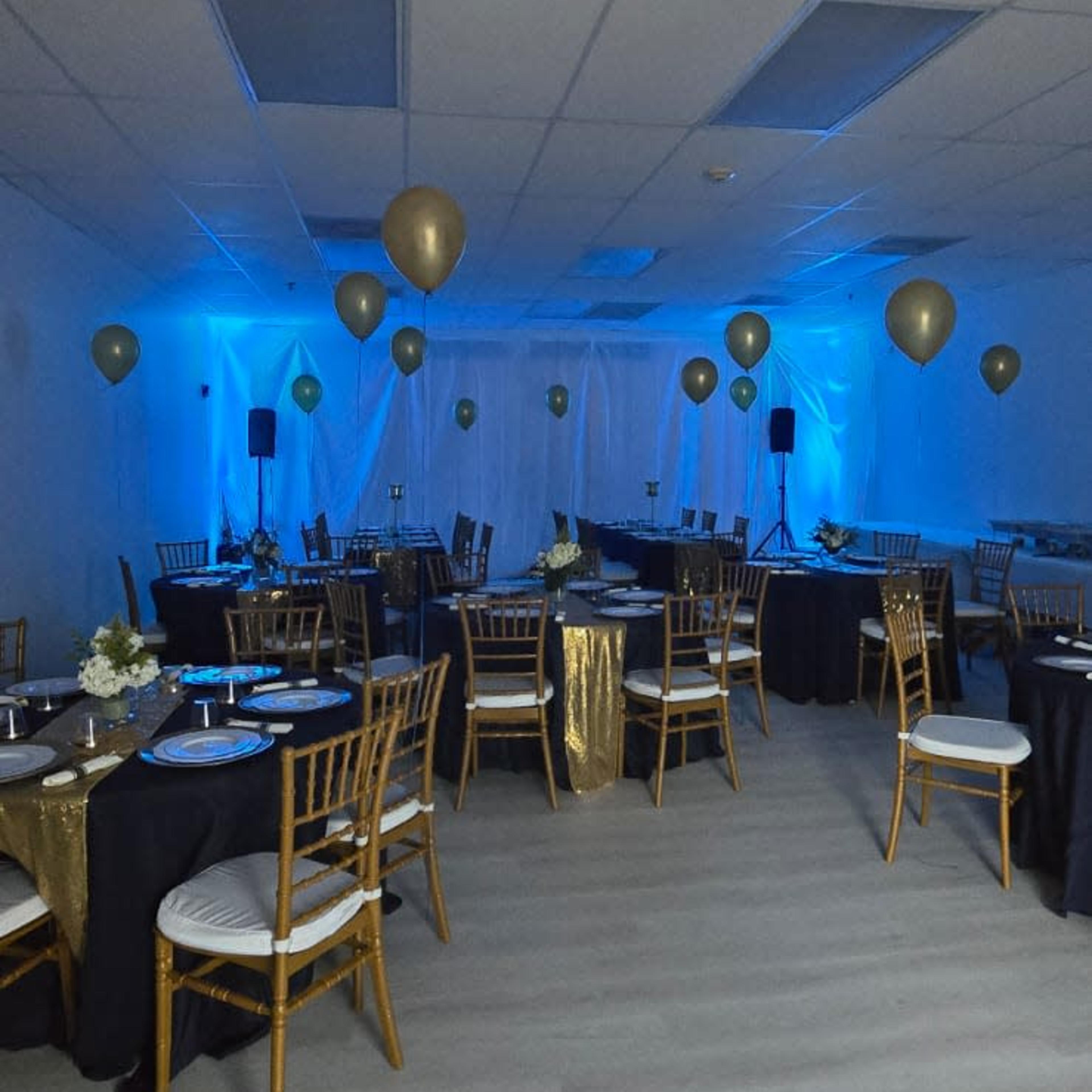 A banquet room is arranged with black and gold tablecloths, chairs, and balloons, featuring blue lighting and a minimalist decor.
