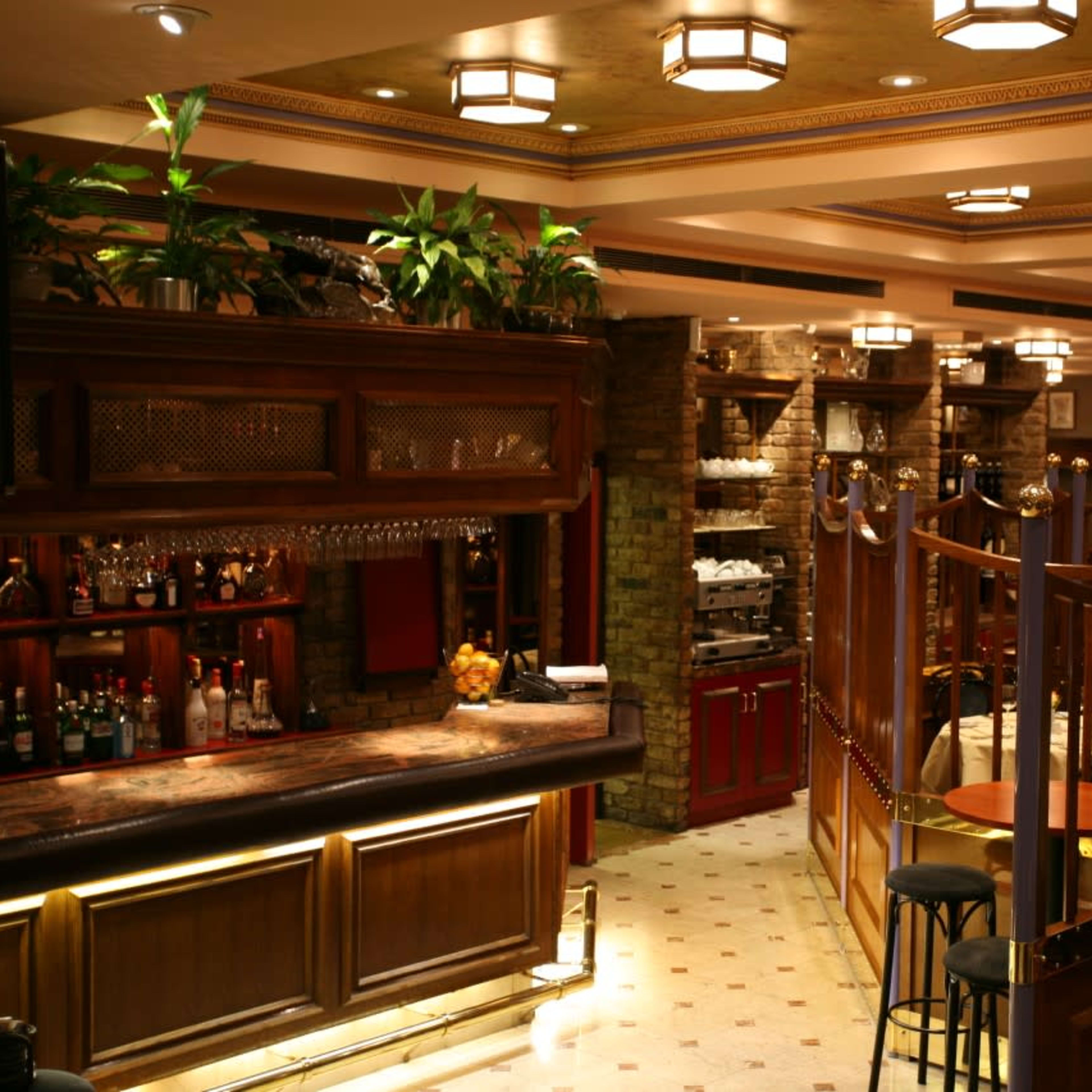 An elegant restaurant interior featuring a wooden bar, various plants, and separate dining areas with round tables and chairs.