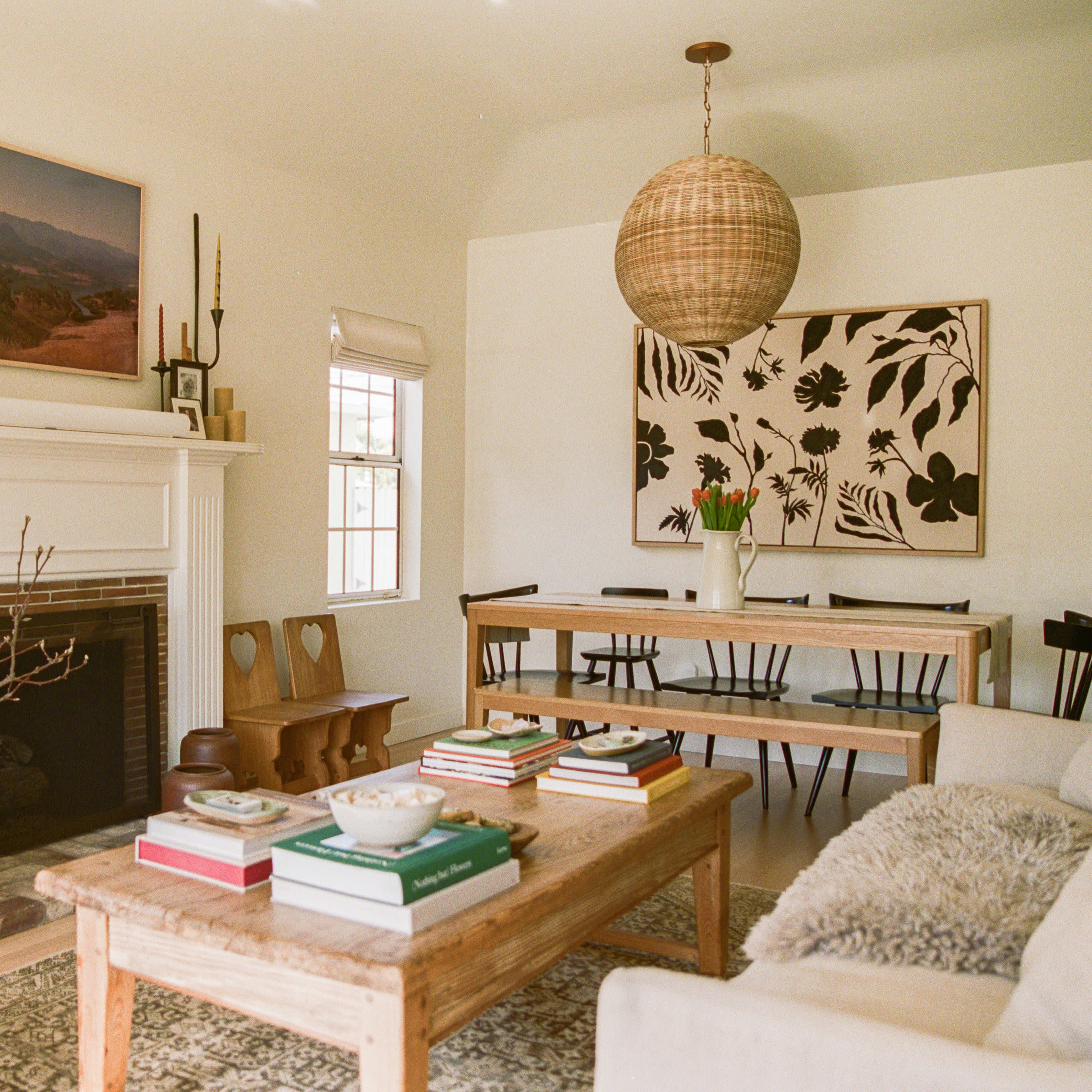 A bright living room featuring a fireplace, a wooden coffee table with stacked books, a dining table, and a large wall art piece.
