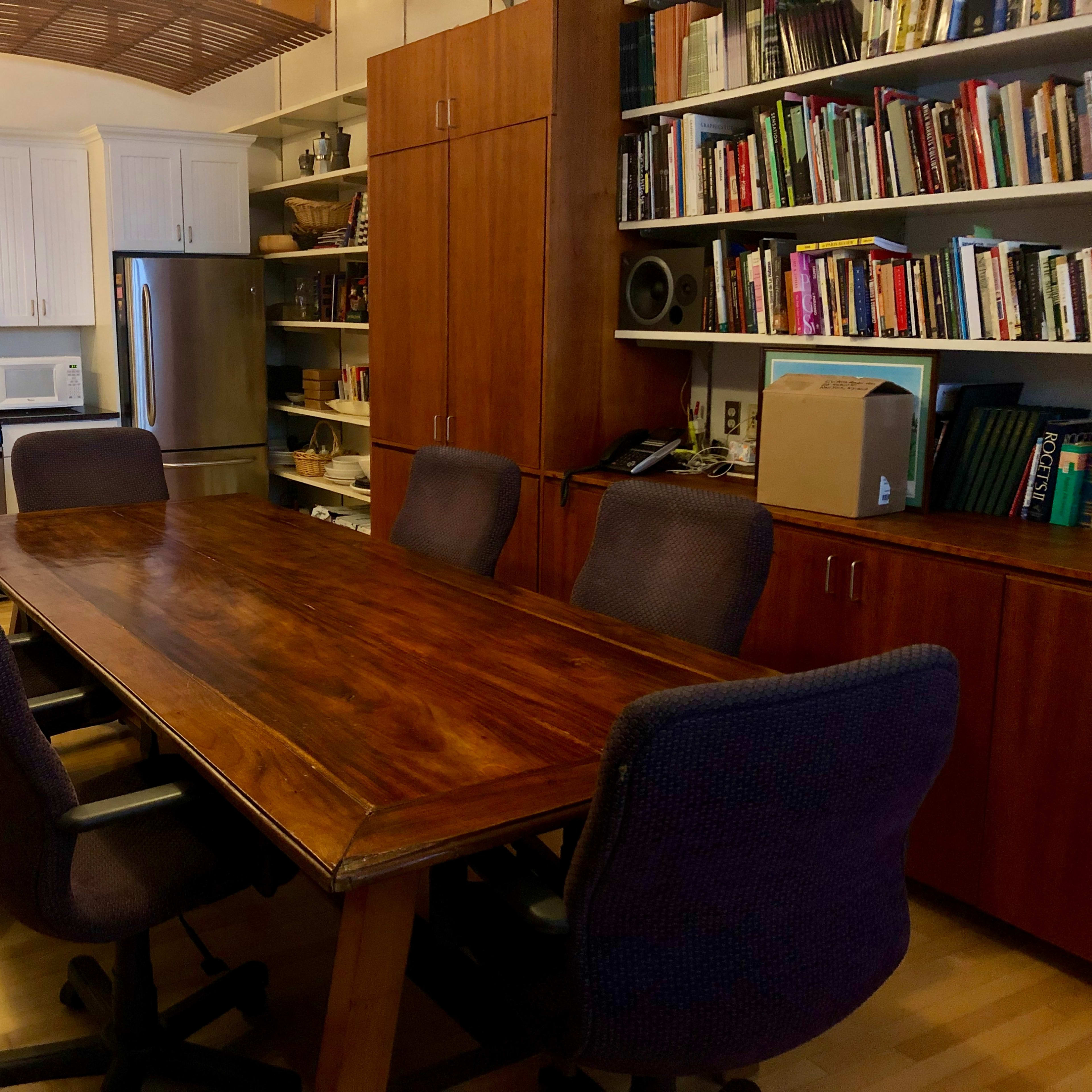 A large wooden conference table surrounded by five ergonomic chairs, situated in a kitchen-like space with a modern kitchen area and bookshelves.