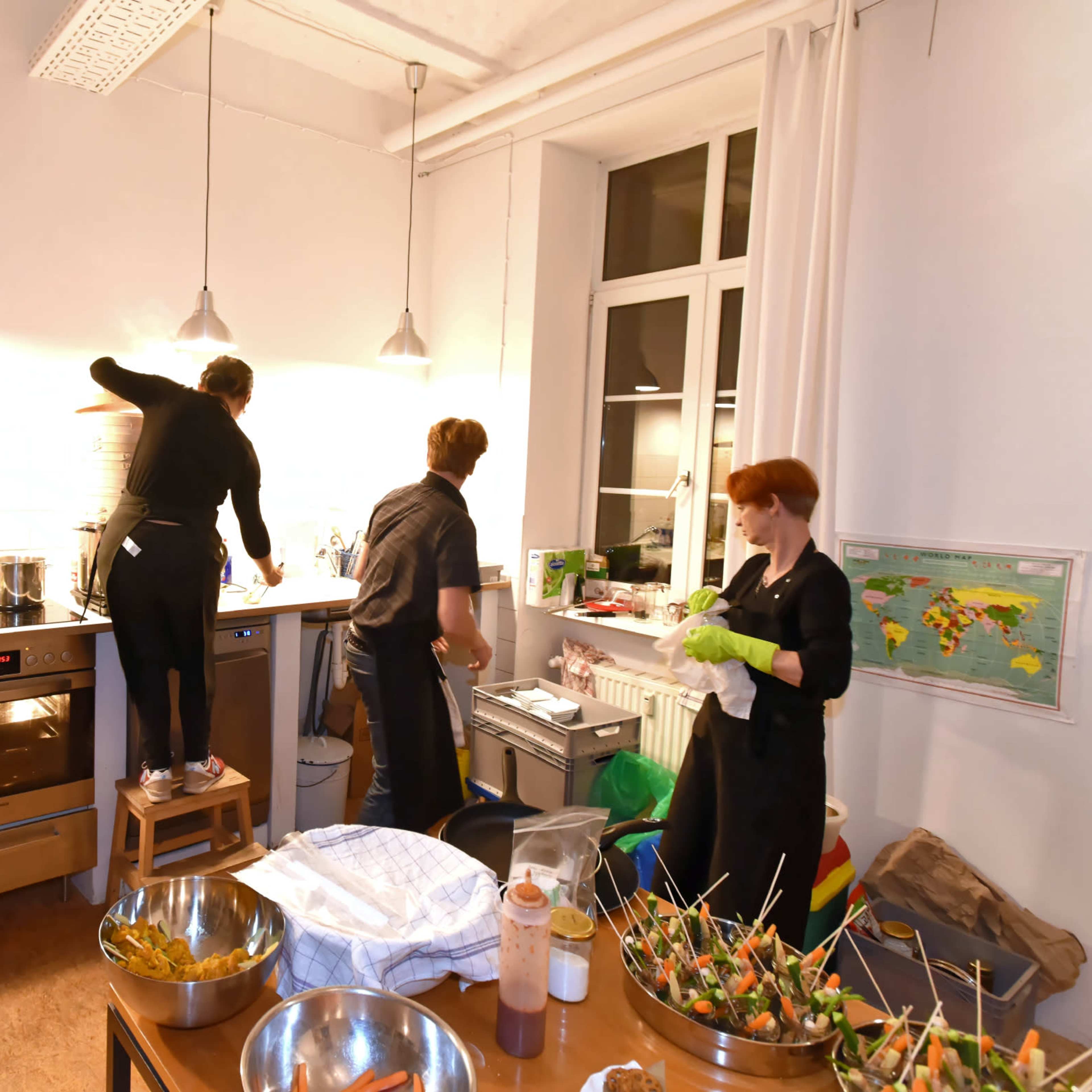Four individuals in black outfits are preparing food and cleaning in a kitchen with modern appliances and large tables.