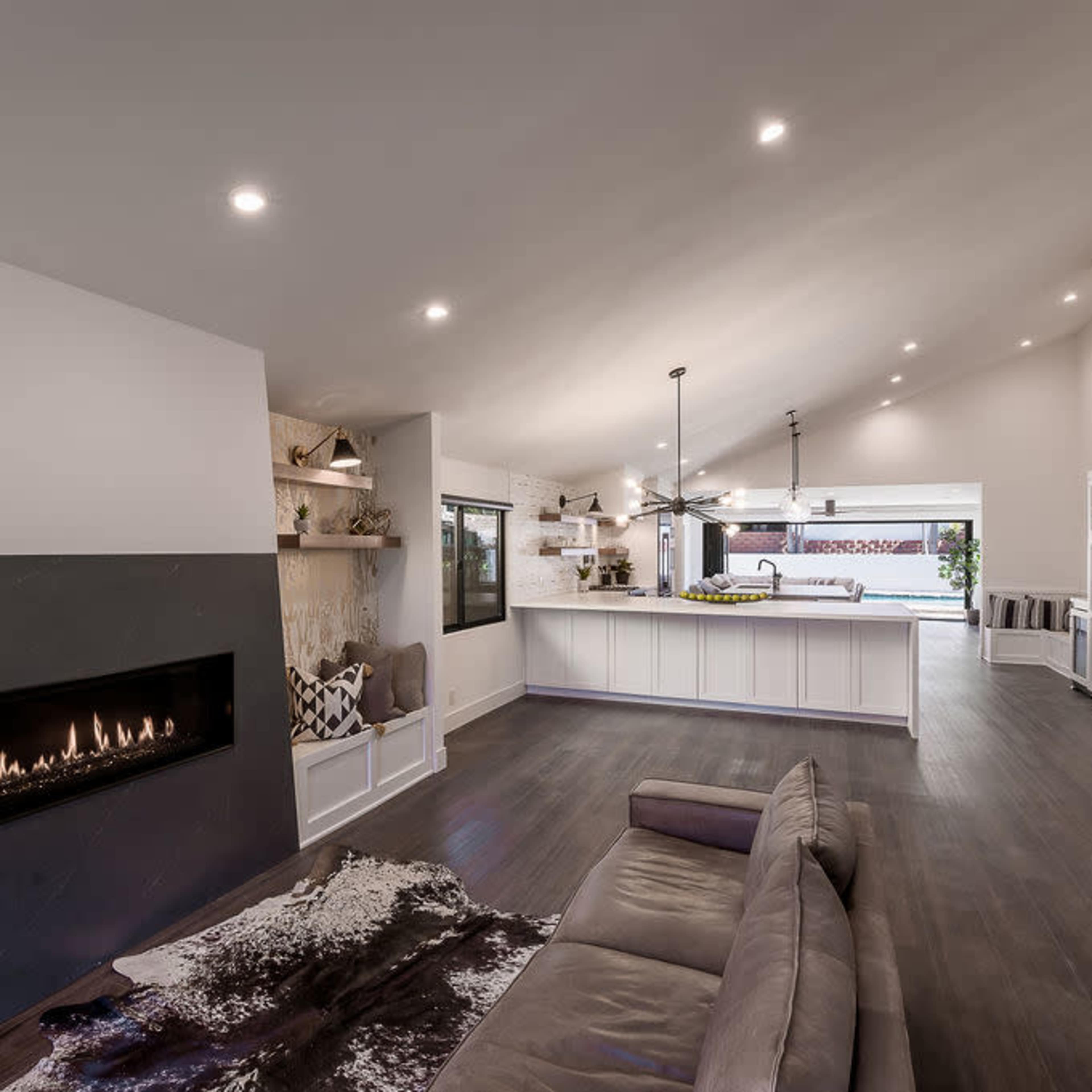 A modern living room with a fireplace, a large open kitchen, and an area with neutral furniture and hardwood flooring.