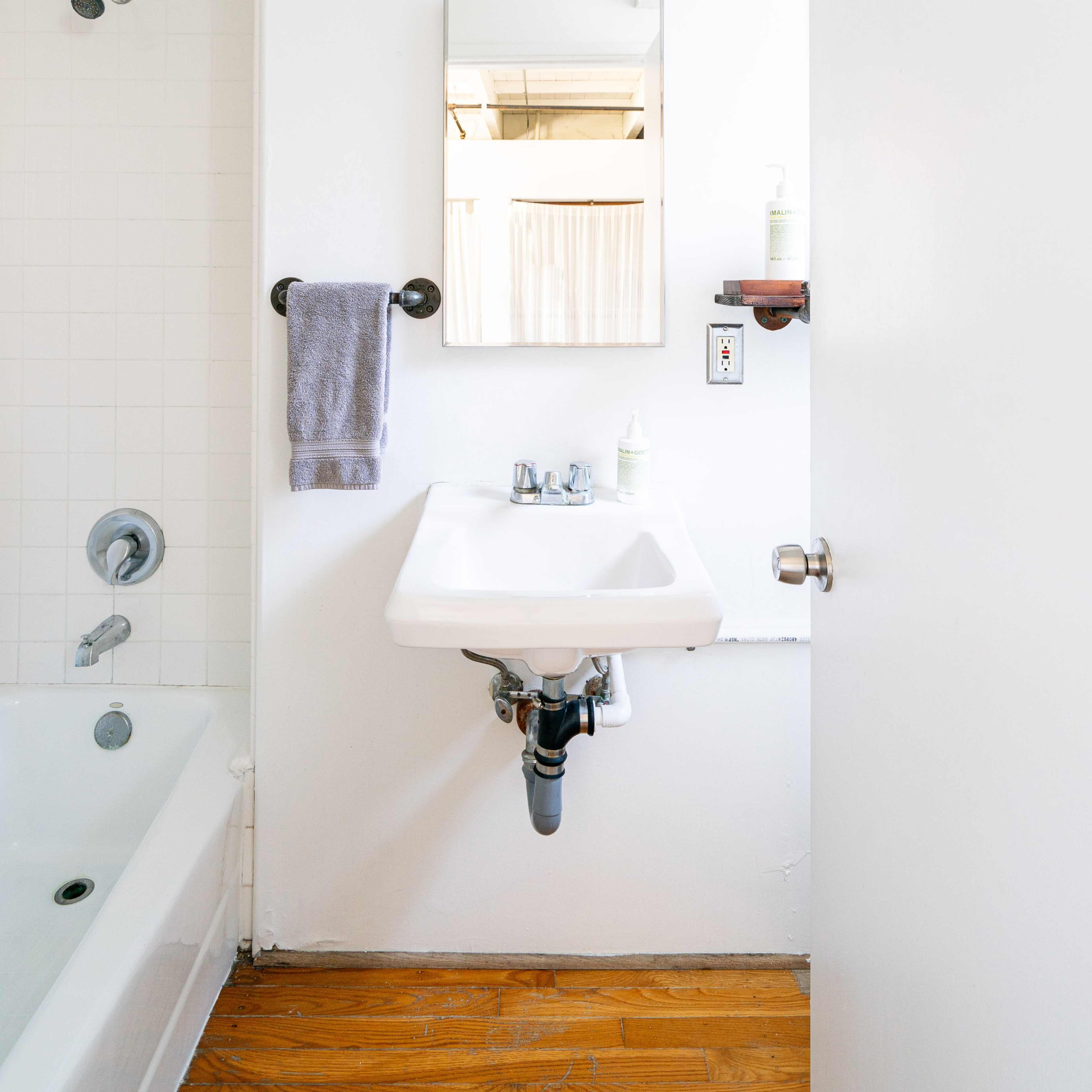 A small, uncluttered bathroom with a white sink, a mirror above it, a towel hanging on the wall, and a bathtub visible in the background.
