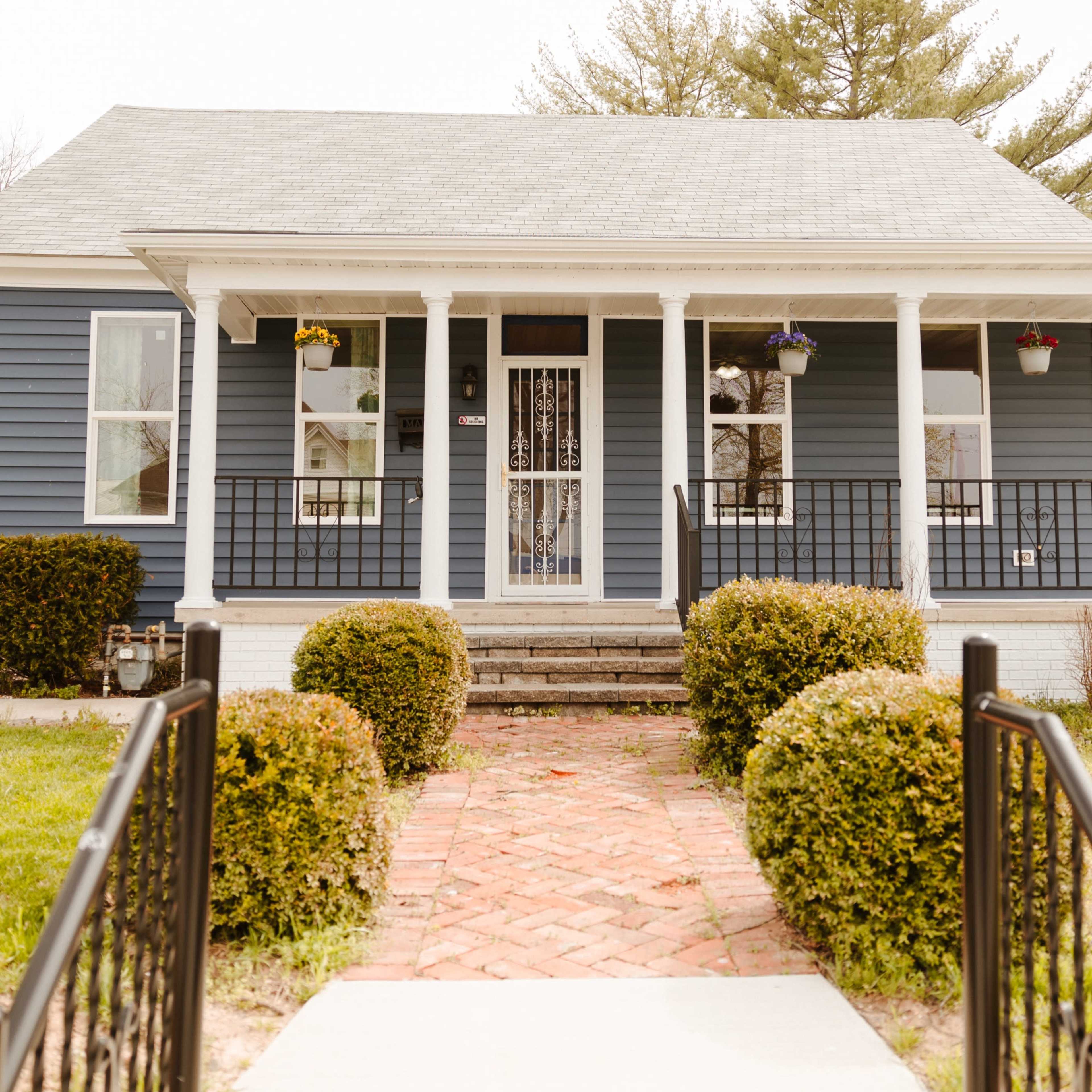 A blue house with white columns and a front porch is surrounded by manicured shrubs and a stone walkway leading to the door.