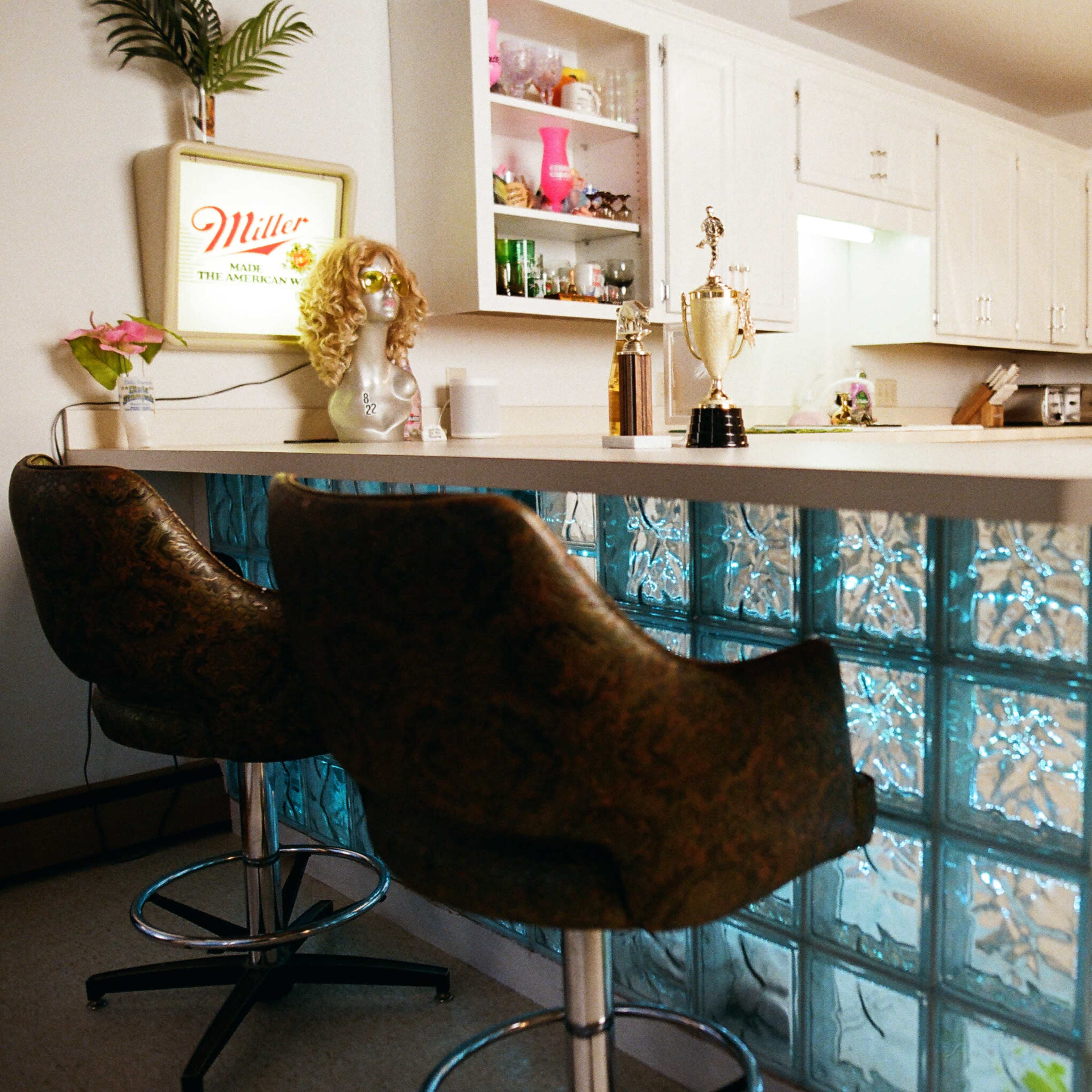 A kitchen with a bar area featuring a glass block design and two upholstered stools, along with pink and yellow decorative items on the countertop and shelves.