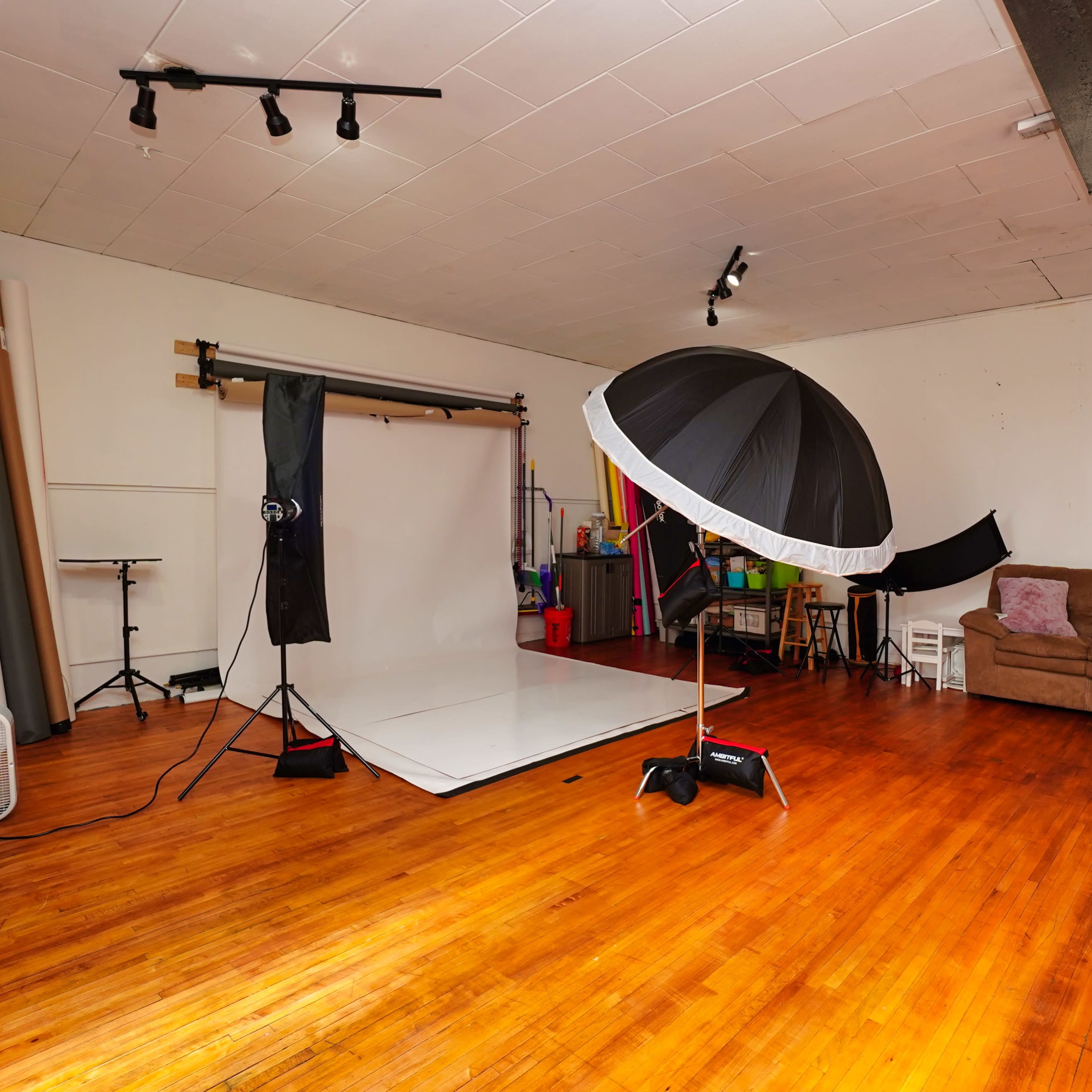 A photography studio with a white backdrop, an umbrella light setup, and a brown couch against the wall.