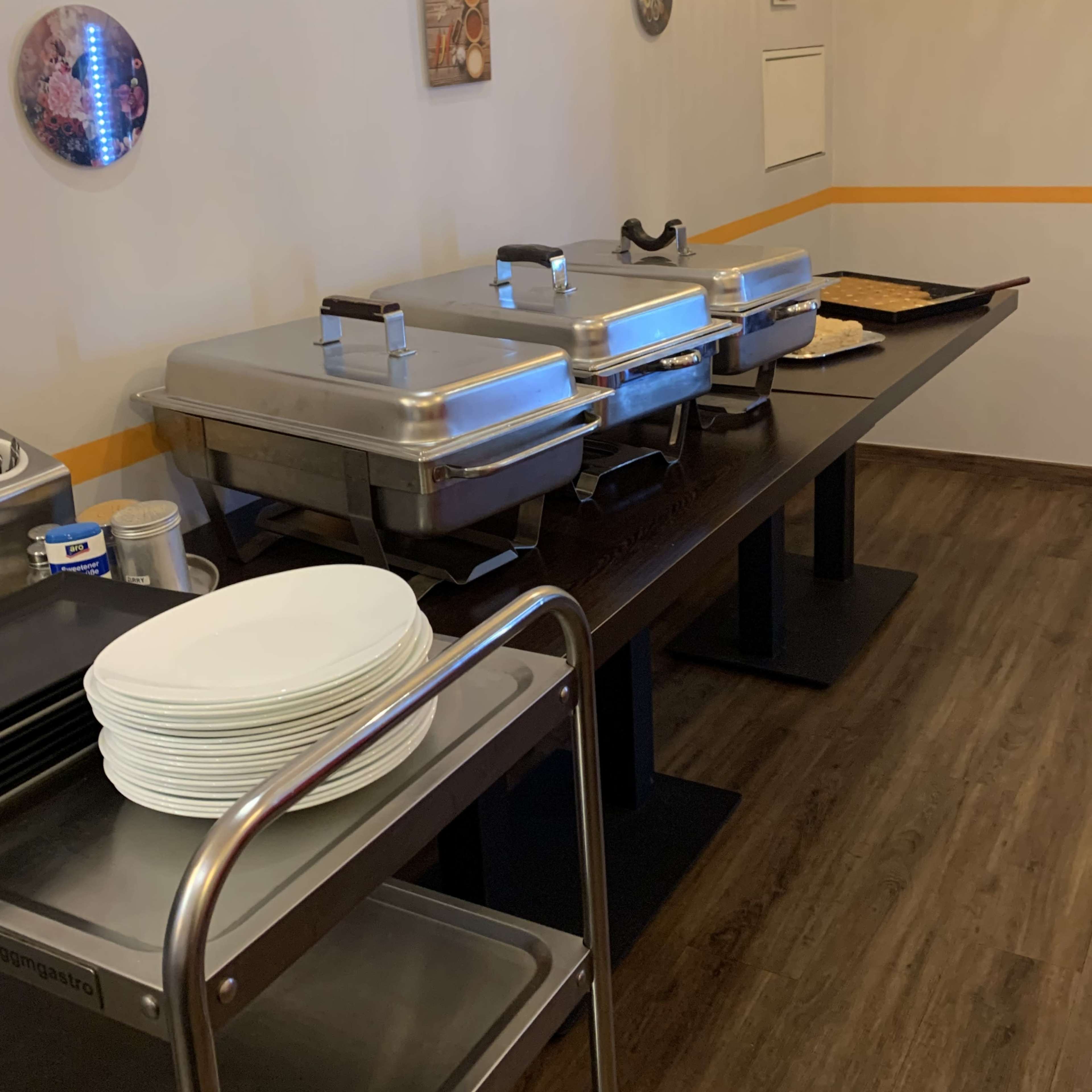 A buffet setup features two stainless steel food warmers and a stack of white plates on a table with wooden floors.