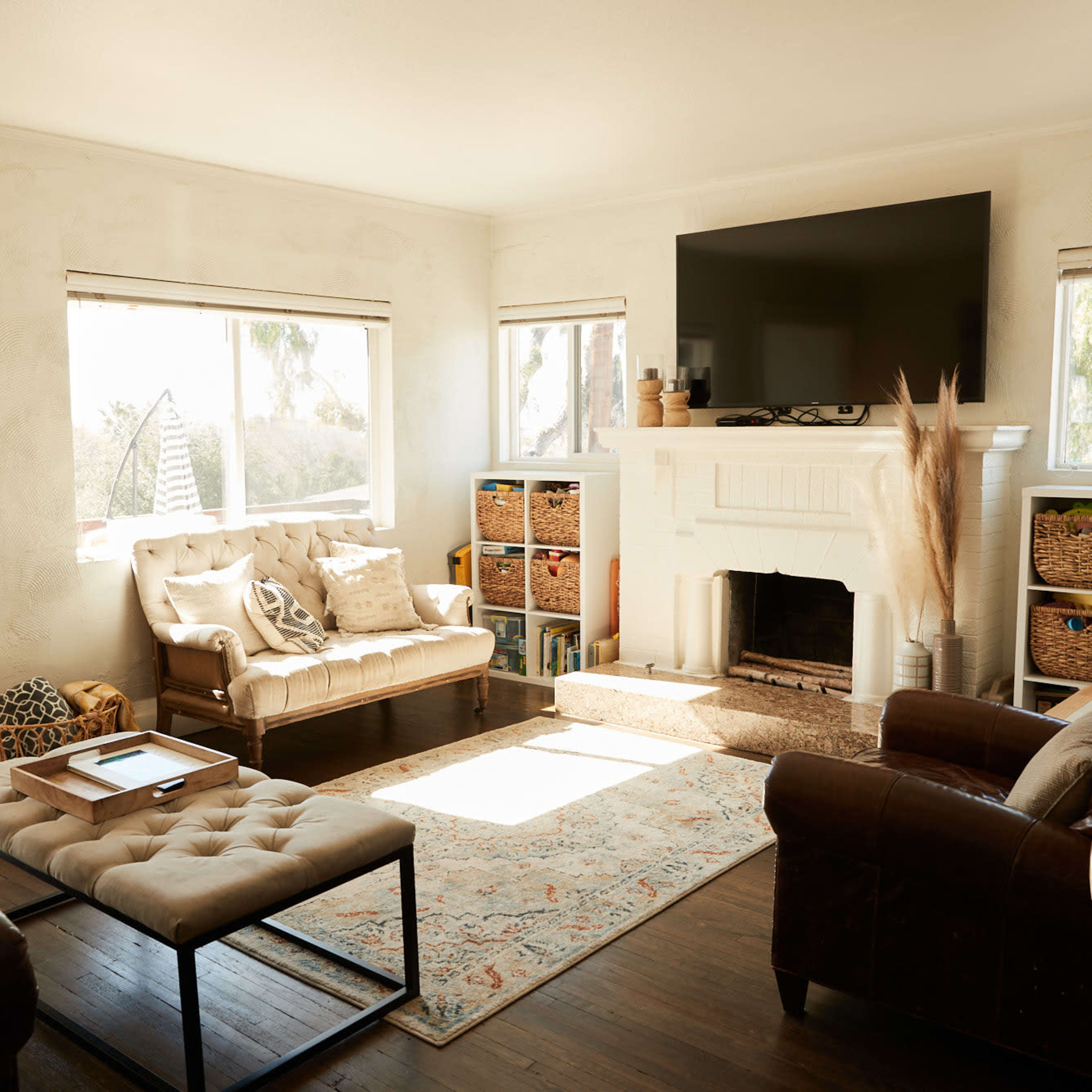 A living room features two sofas, a coffee table, a fireplace with a large television above it, and shelves filled with wicker baskets.