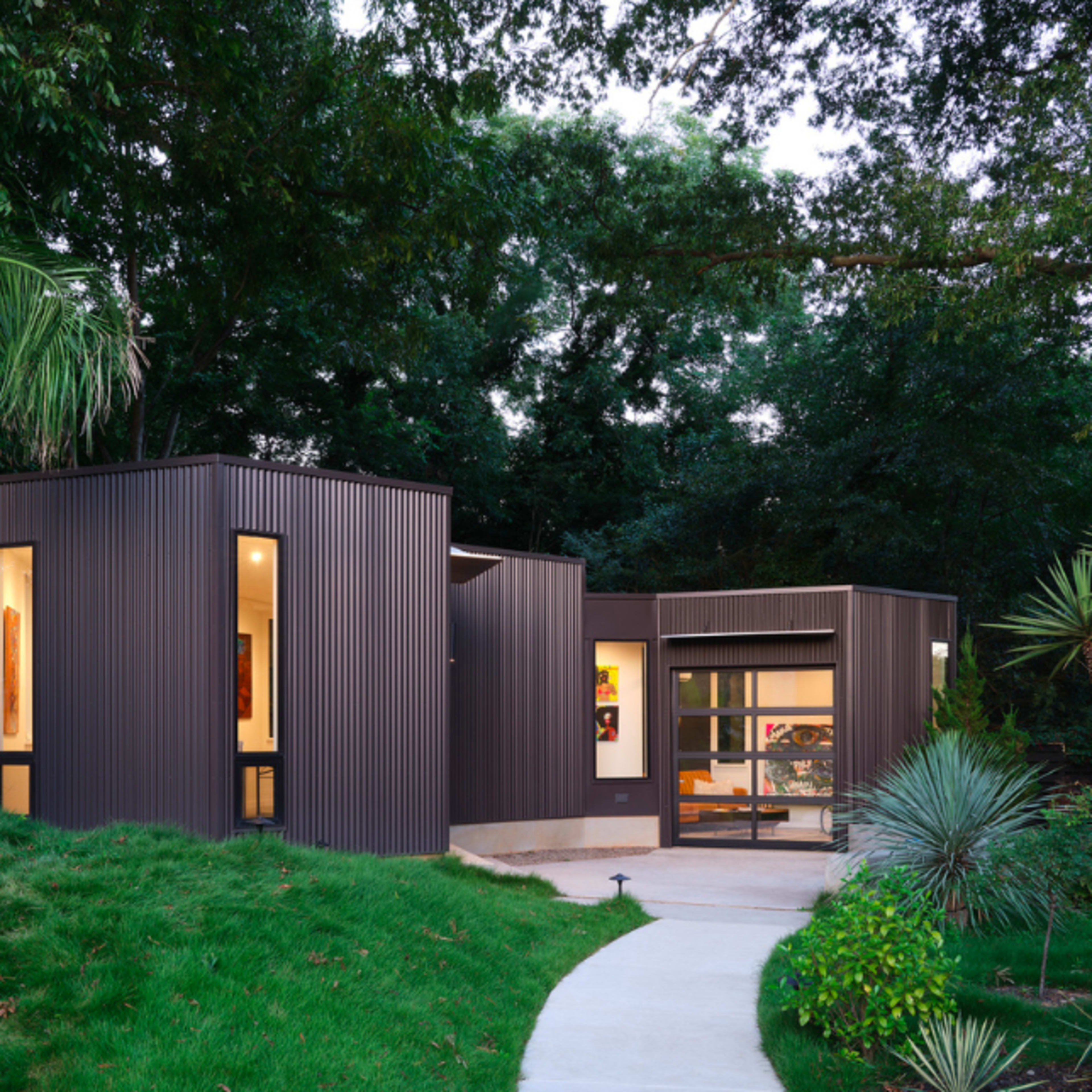 A modern, angular house with dark metal siding is surrounded by lush greenery and features large windows.