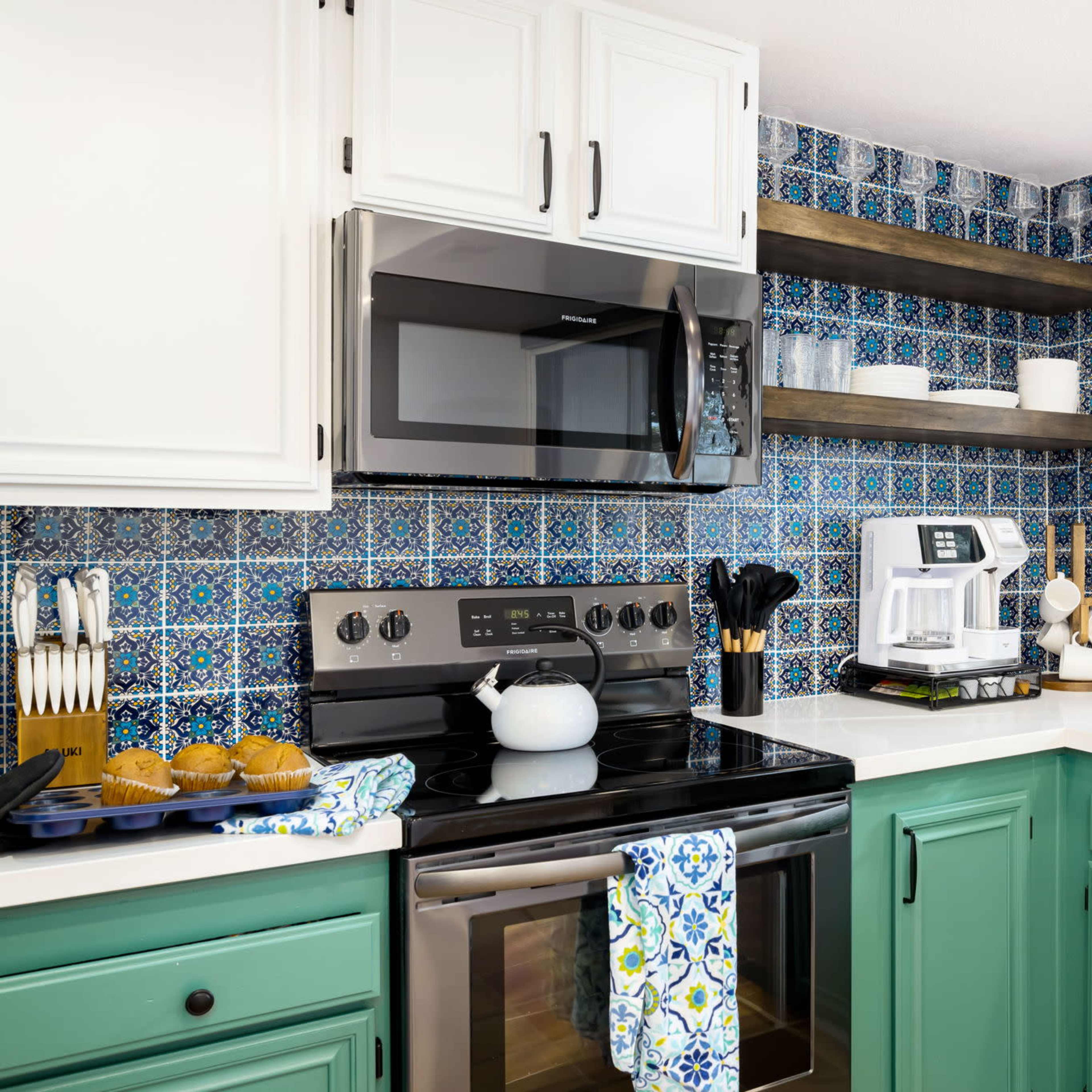 A modern kitchen with teal cabinets, dark countertops, a microwave above the stove, and a window displaying an outdoor view.