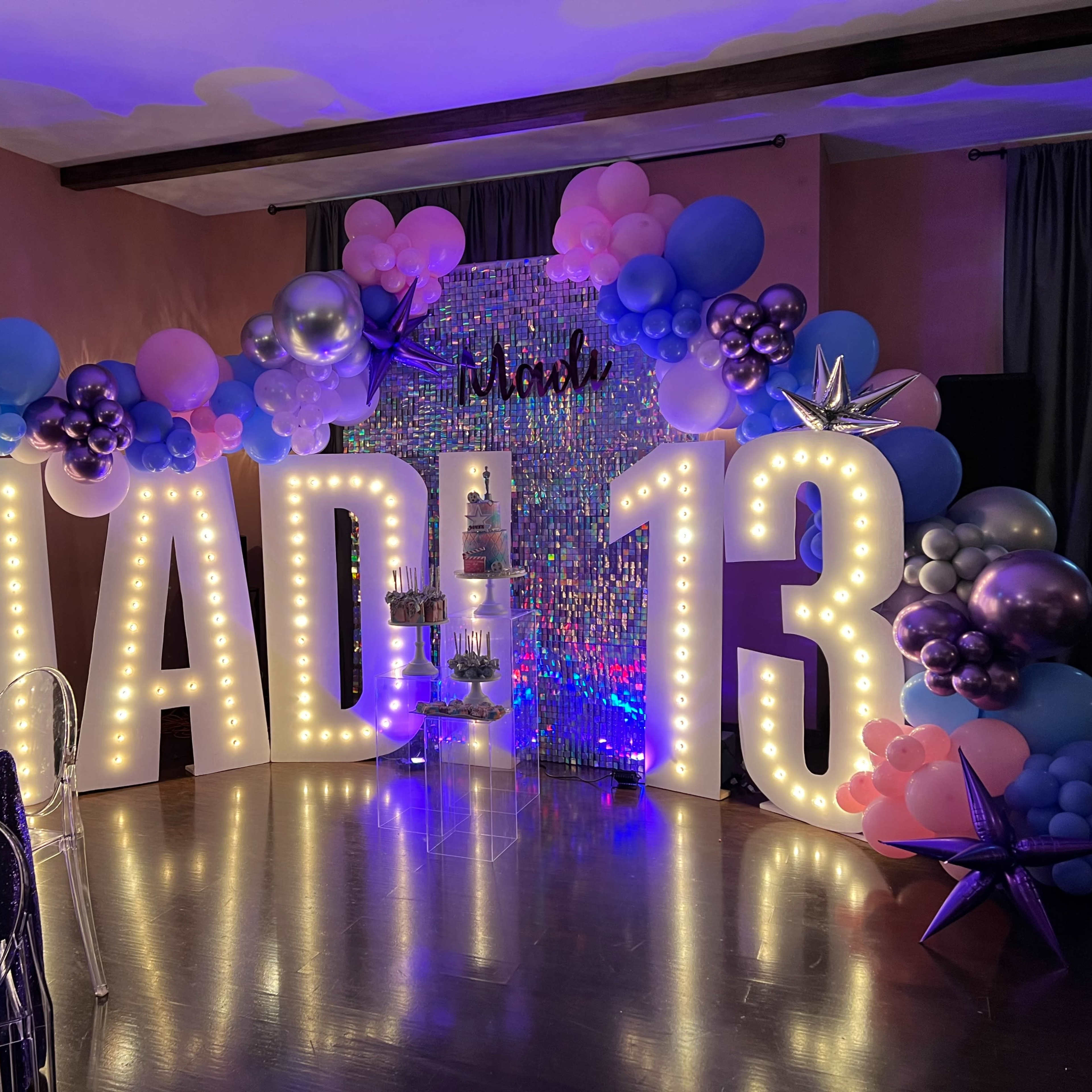 The scene features a decorative setup for a 13th birthday party, highlighted by large illuminated letters spelling "MAD" and "13," surrounded by colorful balloons and a shimmering backdrop.