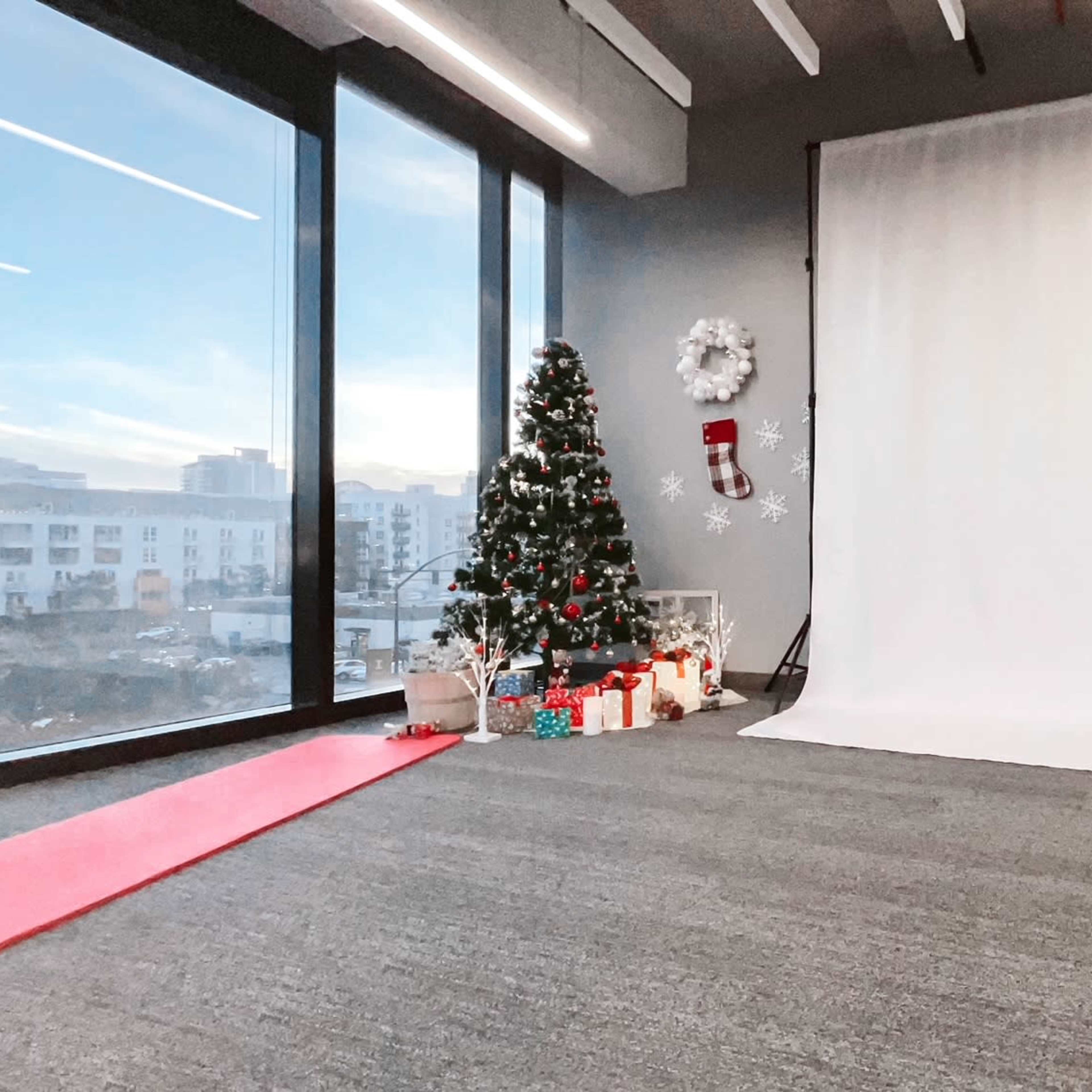 A modern room features a large white backdrop, a Christmas tree with decorations, and gifts arranged on the floor beside a red mat, with large windows displaying an outdoor view.