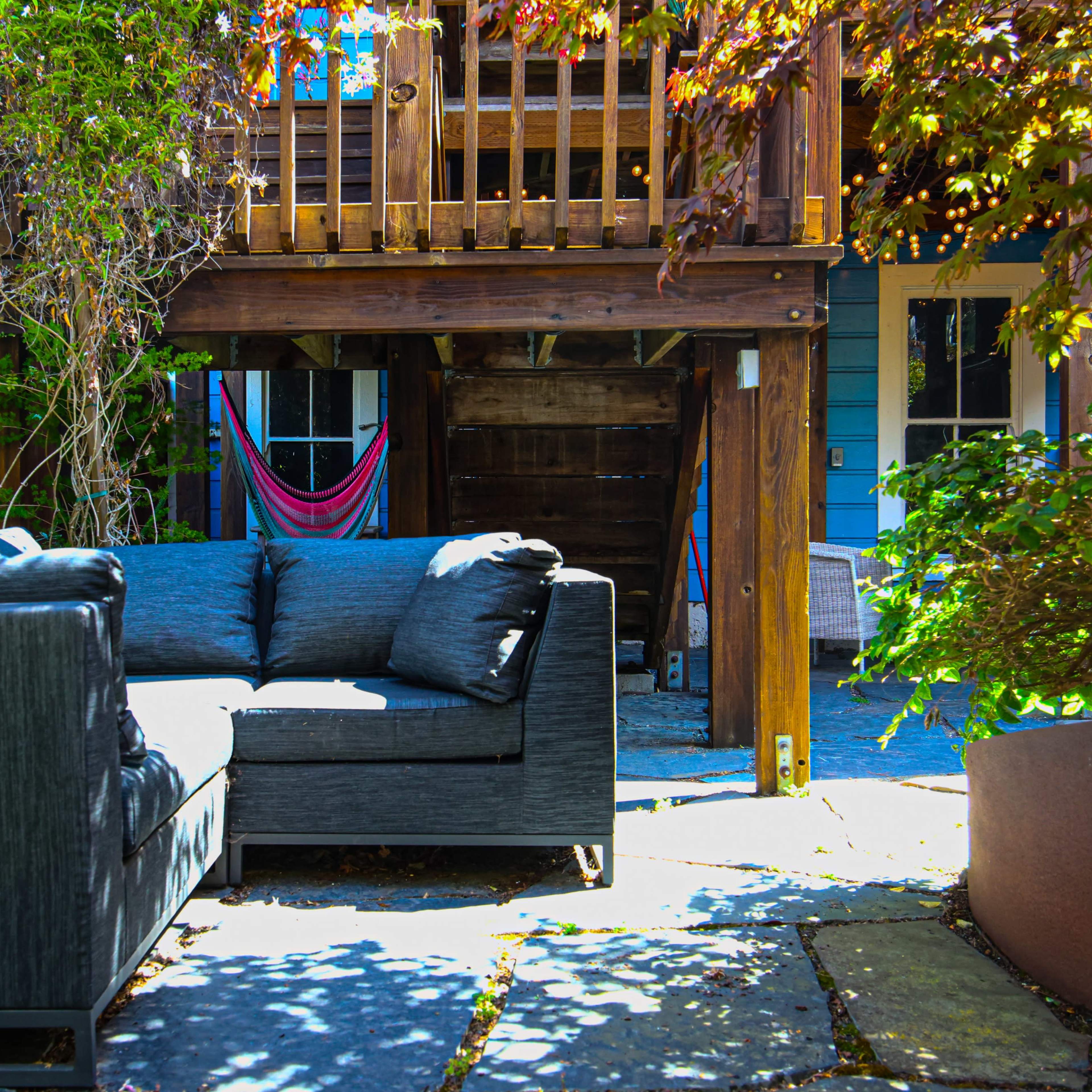 A gray outdoor couch is positioned on a stone patio beneath a wooden deck surrounded by vibrant greenery and a hammock in the background.