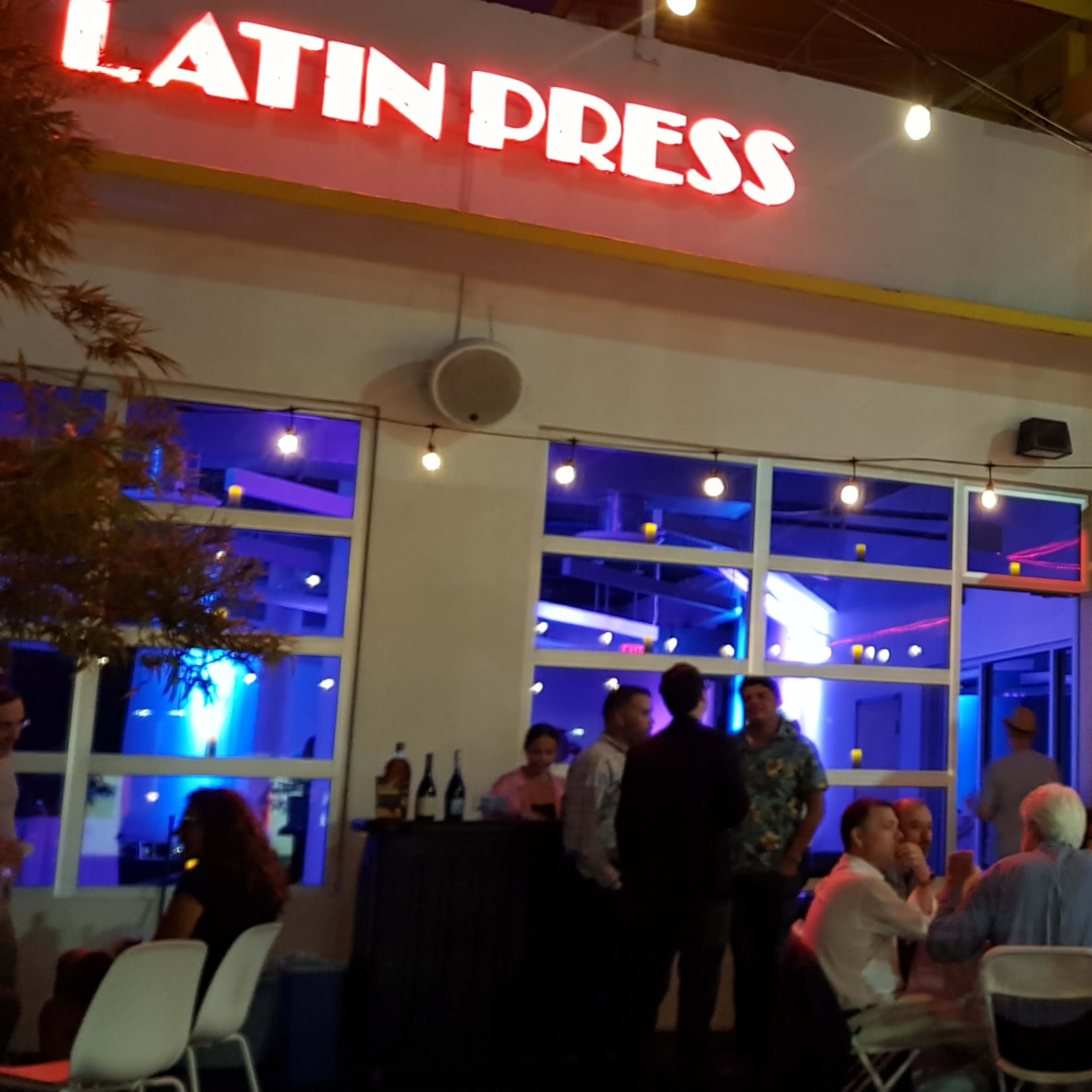 A group of people socialize outside a brightly lit building featuring the words "LATIN PRESS" in neon red lights, with string lights illuminating the area.