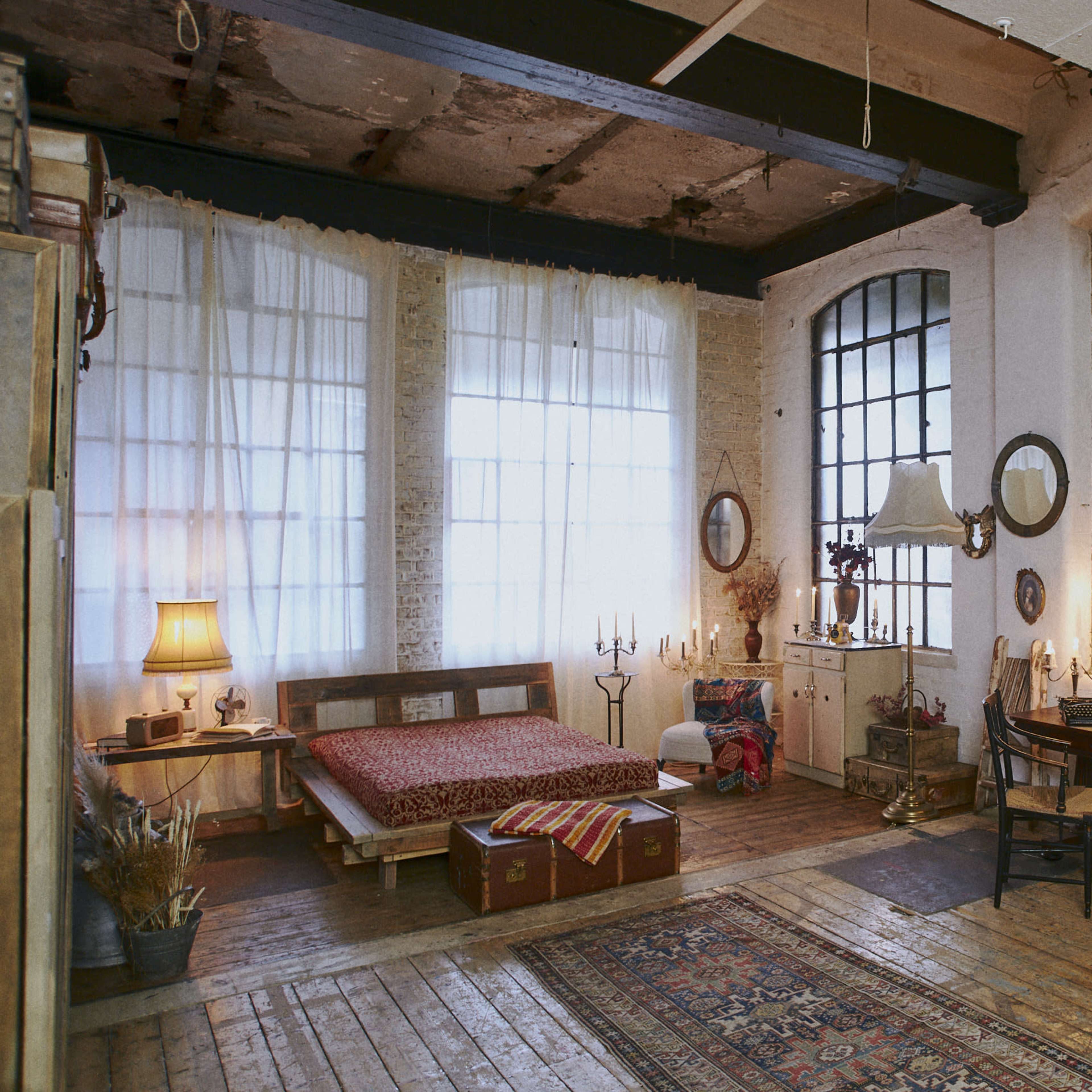 A spacious, rustic bedroom featuring a wooden bed, a desk with a chair, several mirrors, and large windows allowing natural light.