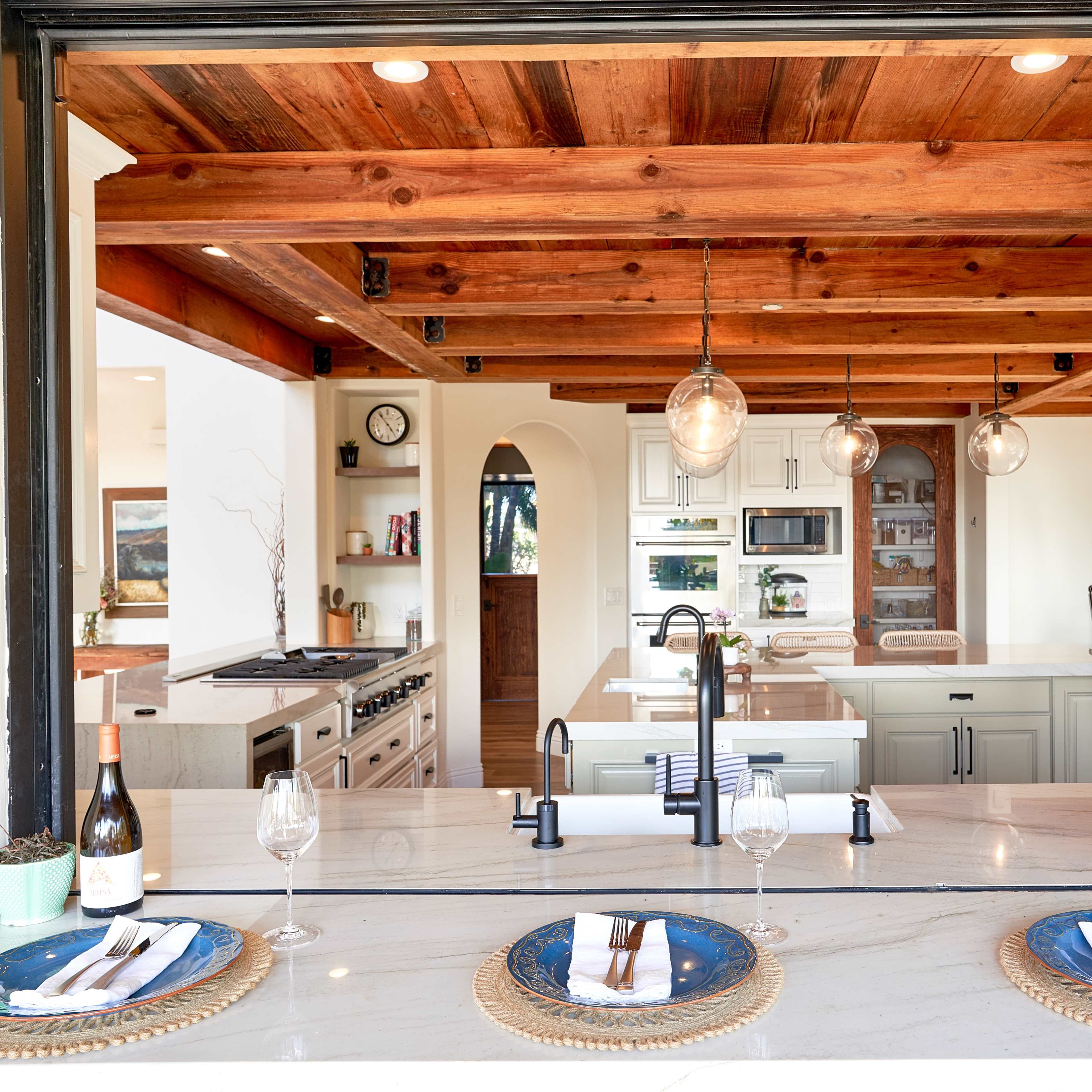 A modern kitchen with wooden beams features an open window overlooking a dining area set with plates and wine glasses.