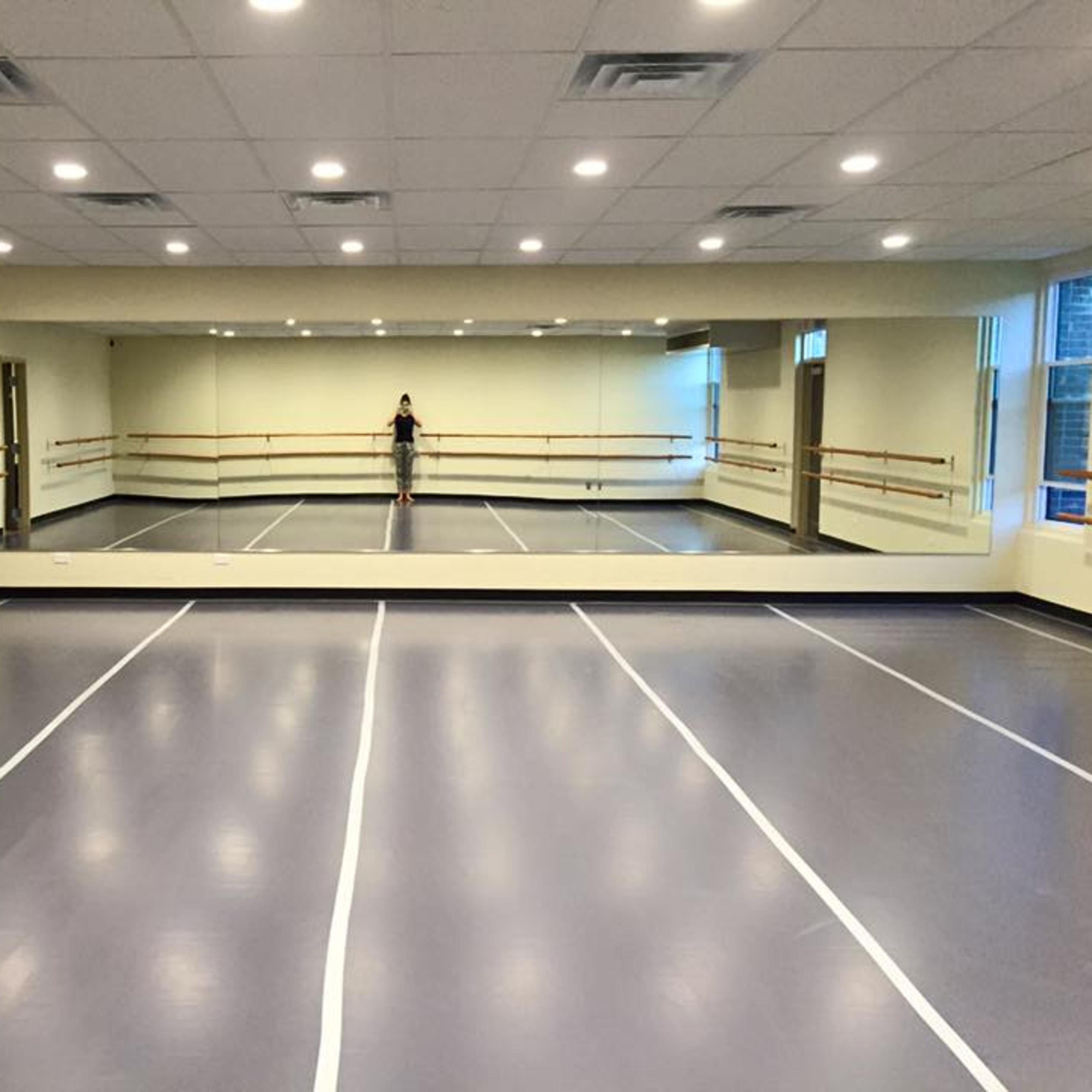 A spacious dance studio features a mirrored wall, barre along the sides, and a gray floor with white marking lines.
